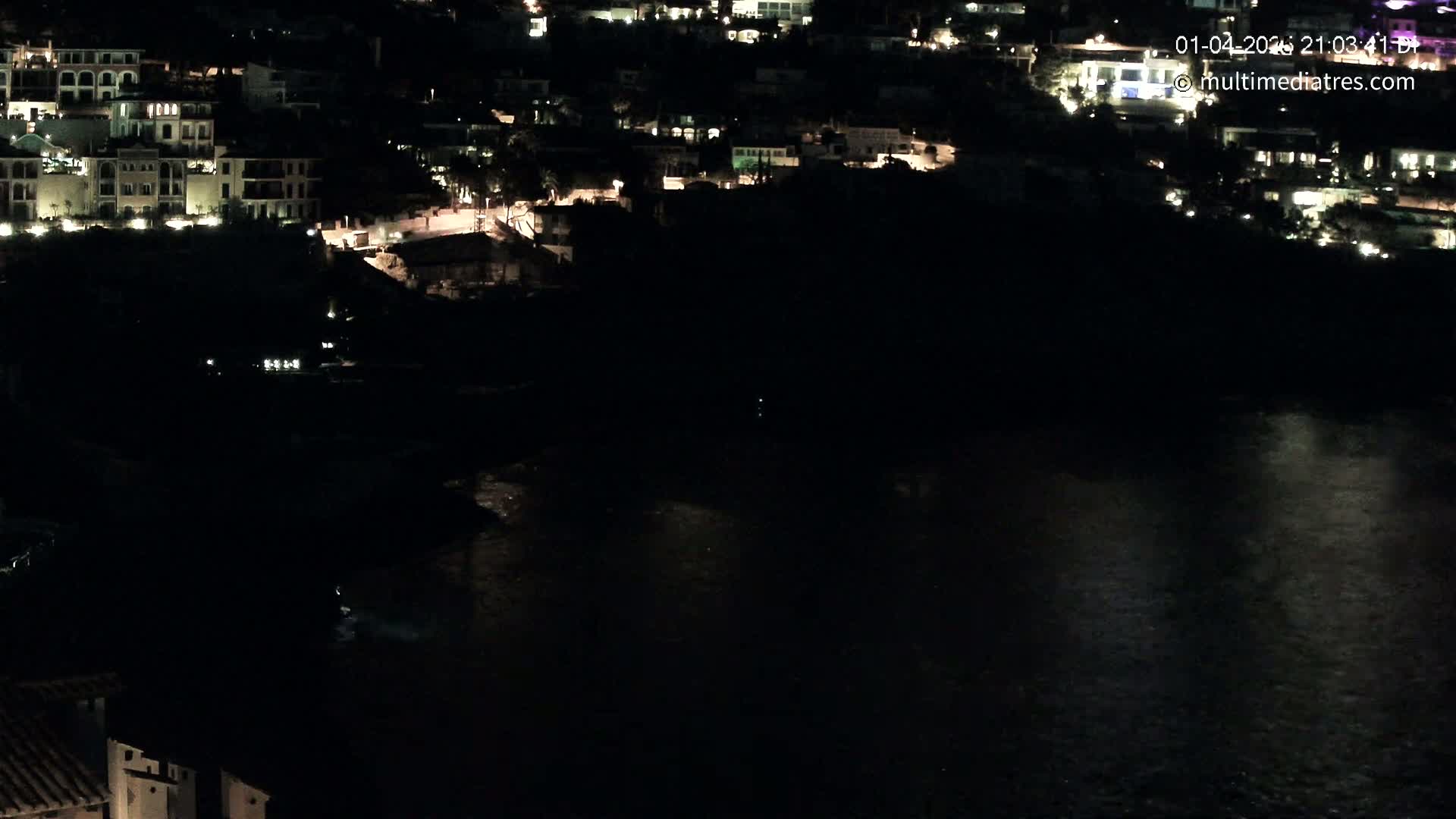 A nighttime, high-angle view shows a dark body of water reflecting a few lights from buildings situated on a hillside.