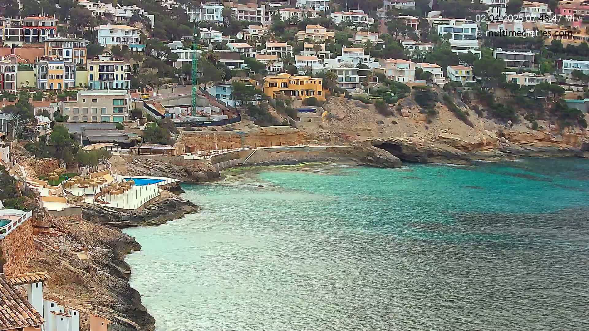 A rocky coastline with a small swimming pool and many houses on a hillside overlooking a teal ocean.
