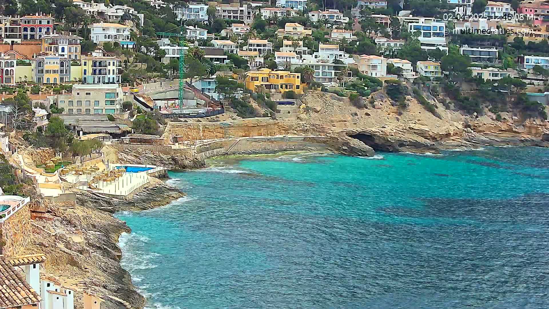 A coastal scene shows a rocky shoreline with turquoise water, numerous buildings on a hillside, and a swimming pool near the water's edge.