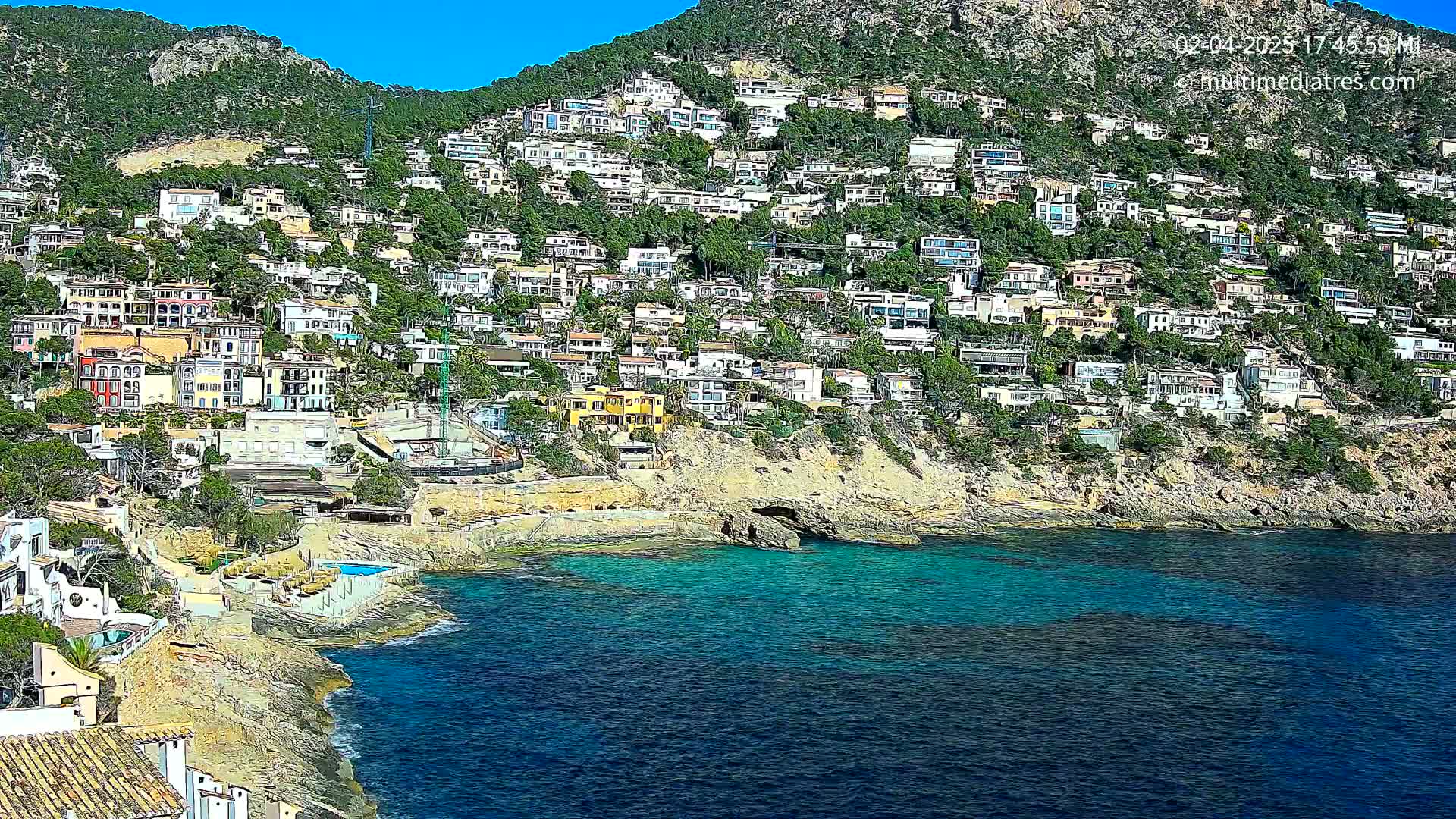 A hillside densely covered with houses slopes down to a clear, teal-colored sea on a sunny day.