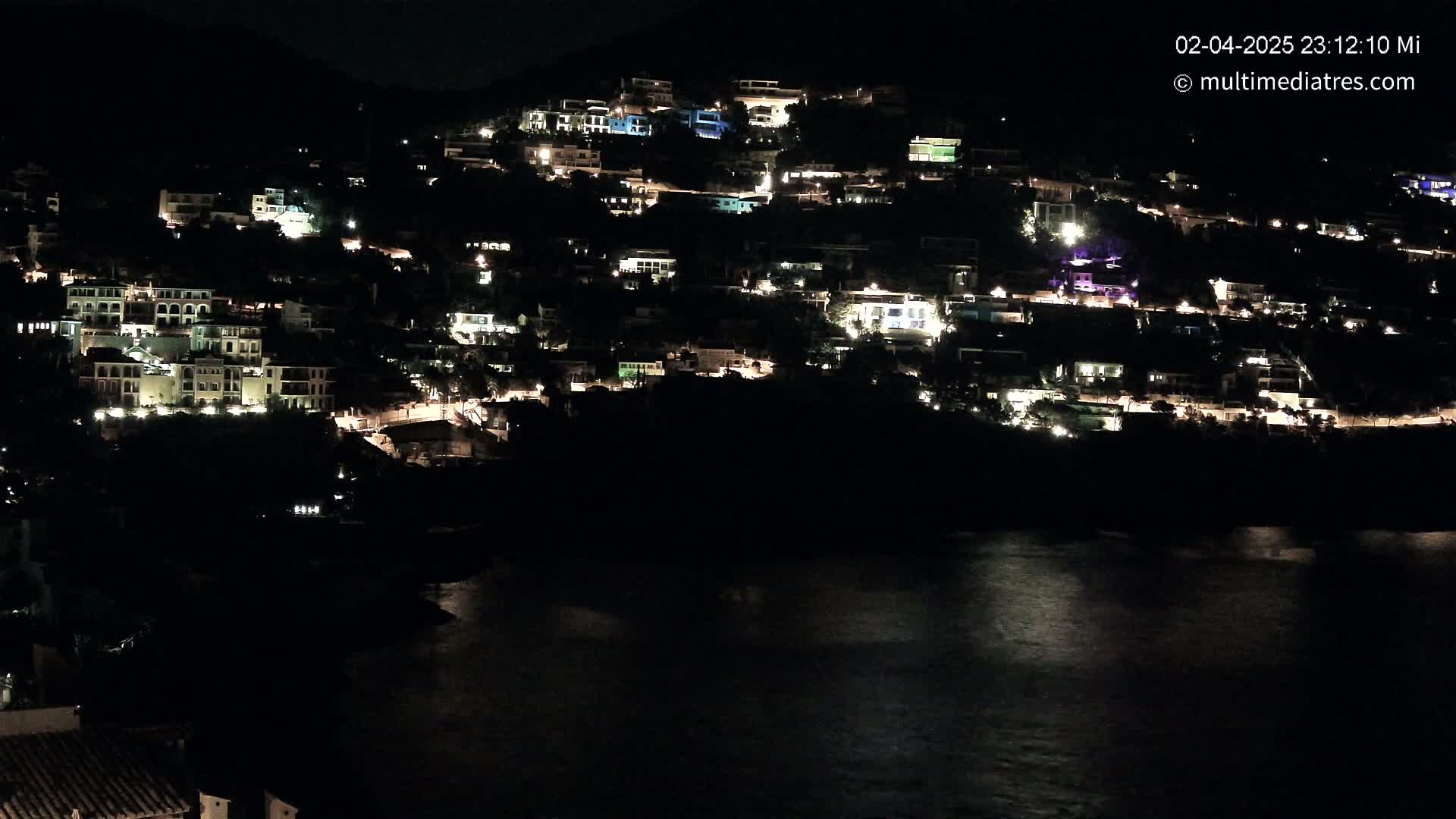 A hillside village is illuminated at night, overlooking a calm body of water.