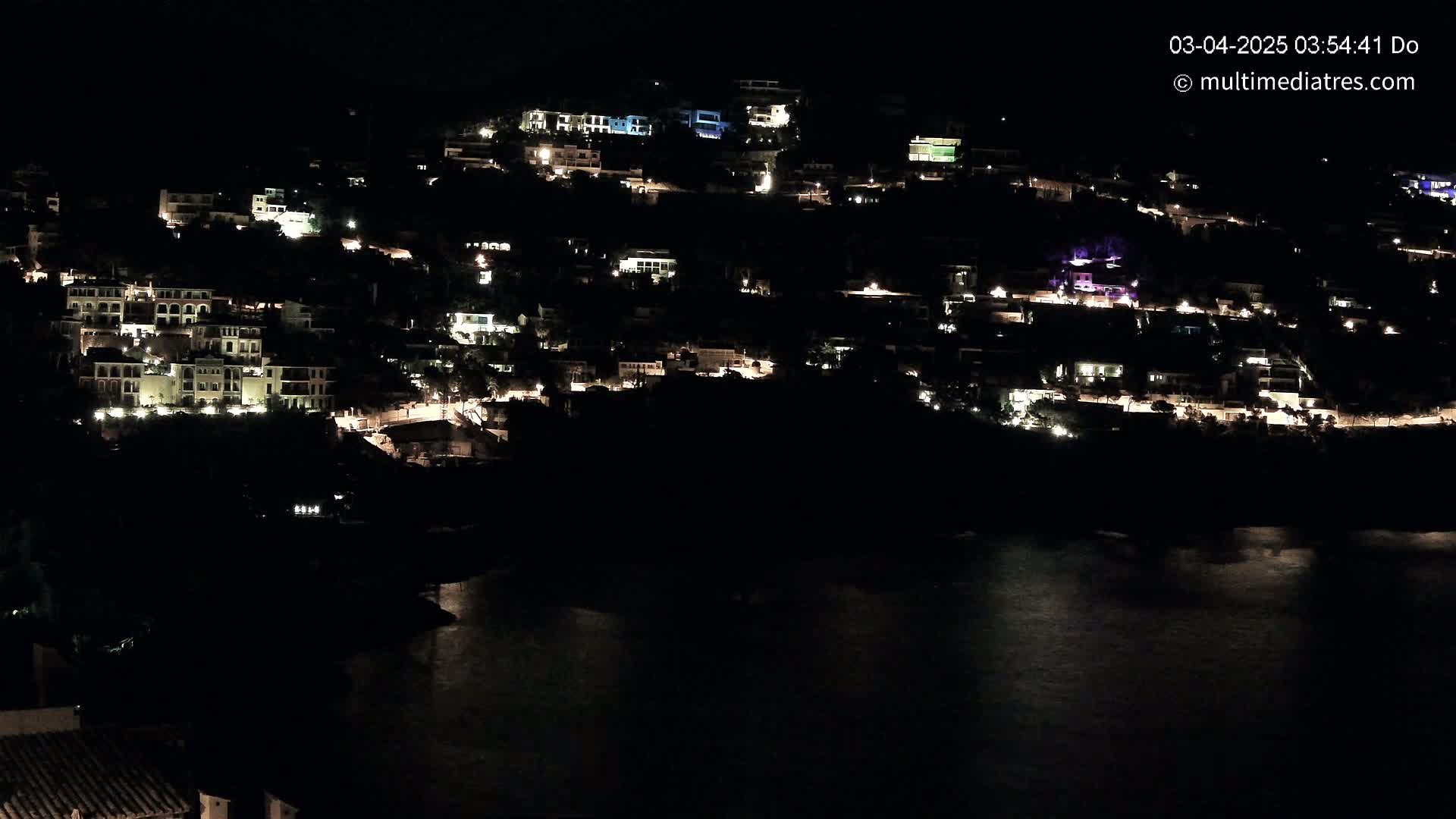 A hillside village is illuminated at night, overlooking a dark body of water.