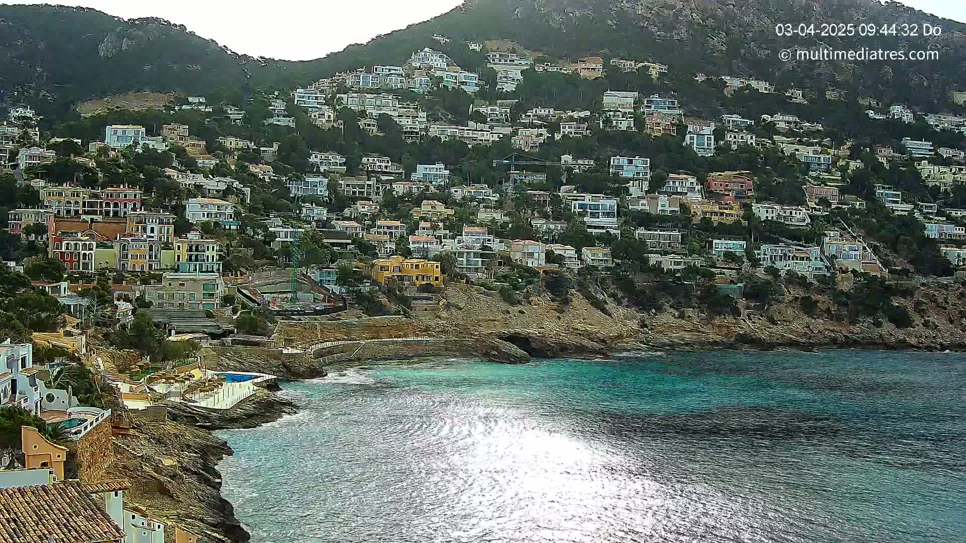 A hillside densely covered with houses slopes down to a calm, sparkling blue sea on a mostly cloudy day.