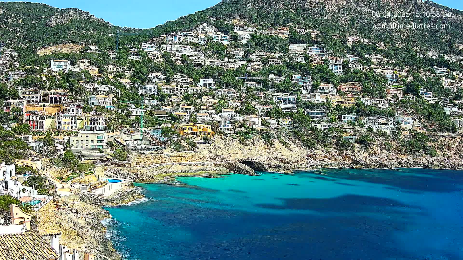 A hillside densely populated with houses overlooks a clear, turquoise bay on a sunny day.