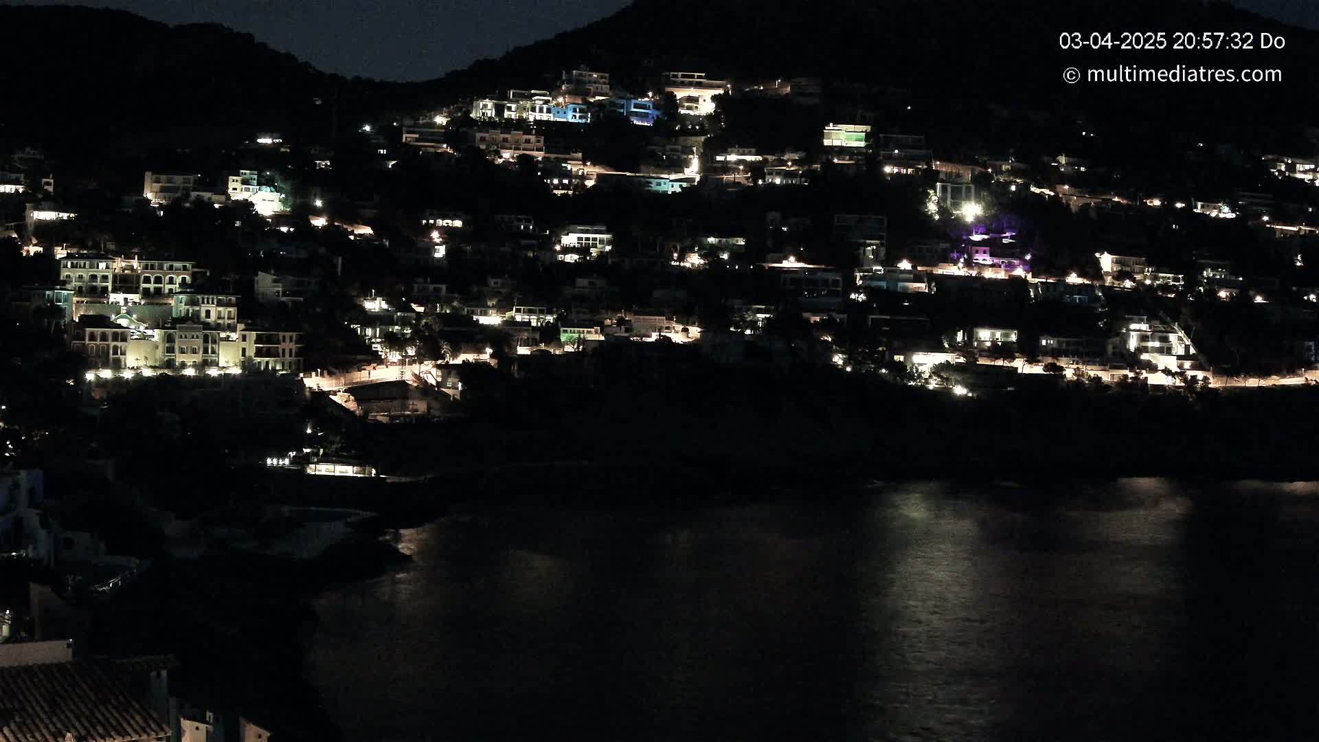 A hillside town at night, illuminated by many lights, sits beside a dark body of water under a clear, dark sky.