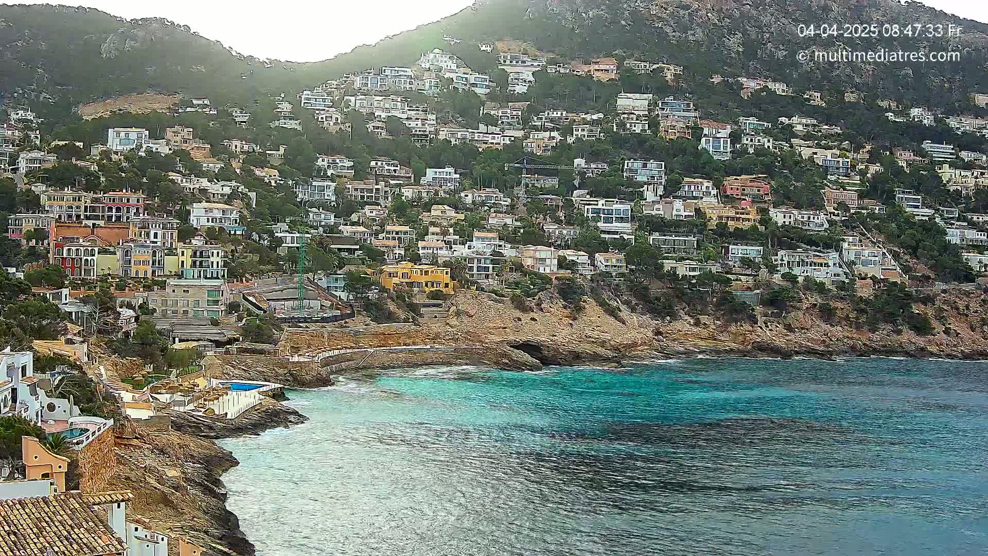 A hillside densely covered with houses overlooks a calm, turquoise bay on an overcast day.