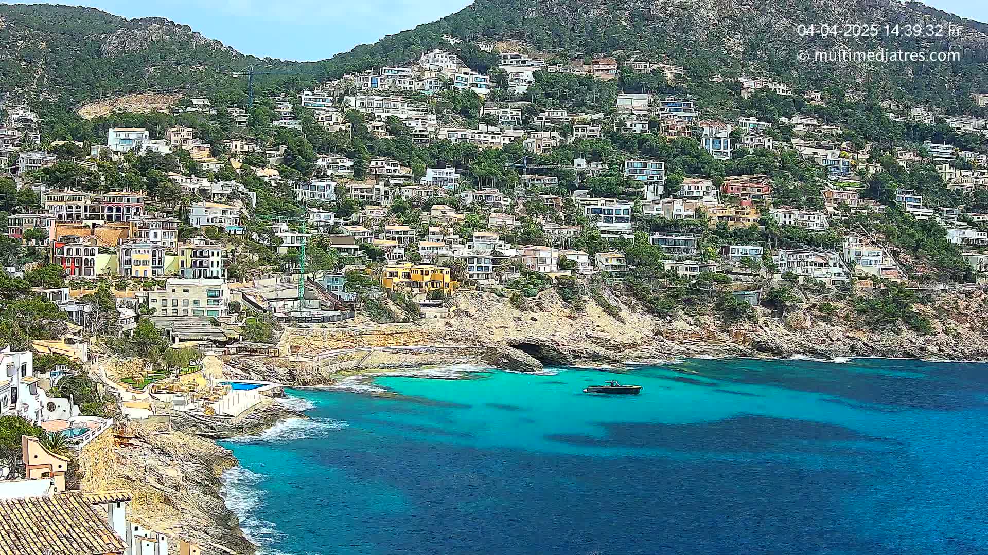 A hillside town of many houses sits above a clear, teal ocean cove on a sunny day.
