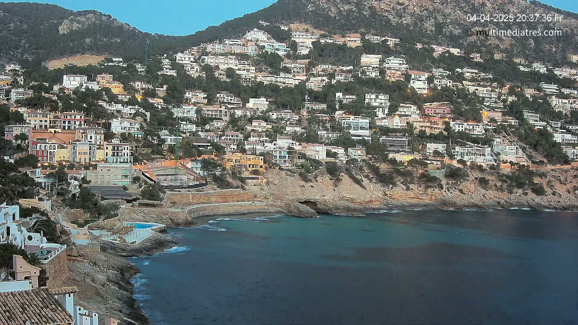 A hillside densely populated with houses overlooks a calm, dark-blue bay under a twilight sky.