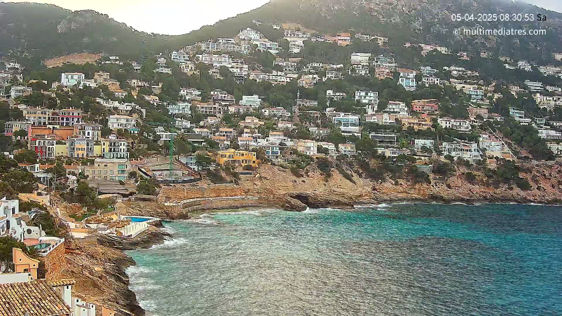 A hillside densely packed with houses overlooks a calm, turquoise ocean under an overcast sky.