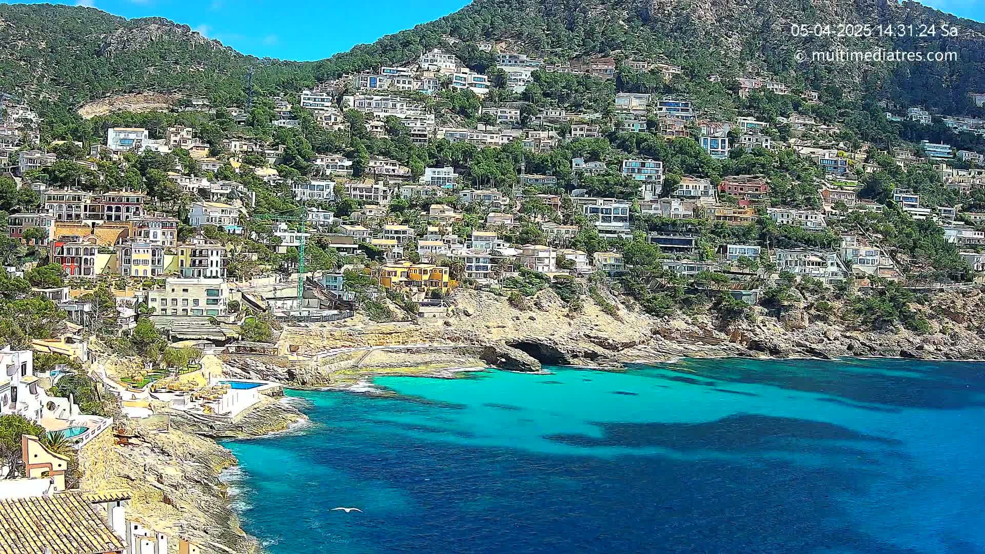 A hillside densely covered with houses overlooks a calm, turquoise cove on a sunny day.