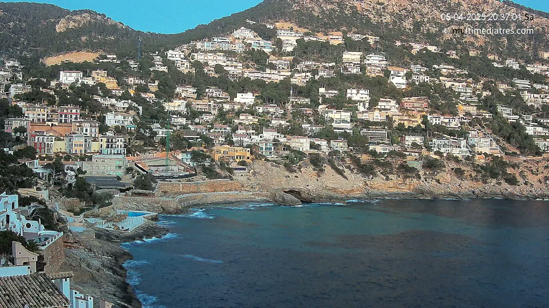A hillside densely populated with houses overlooks a calm, blue ocean under a clear sky.
