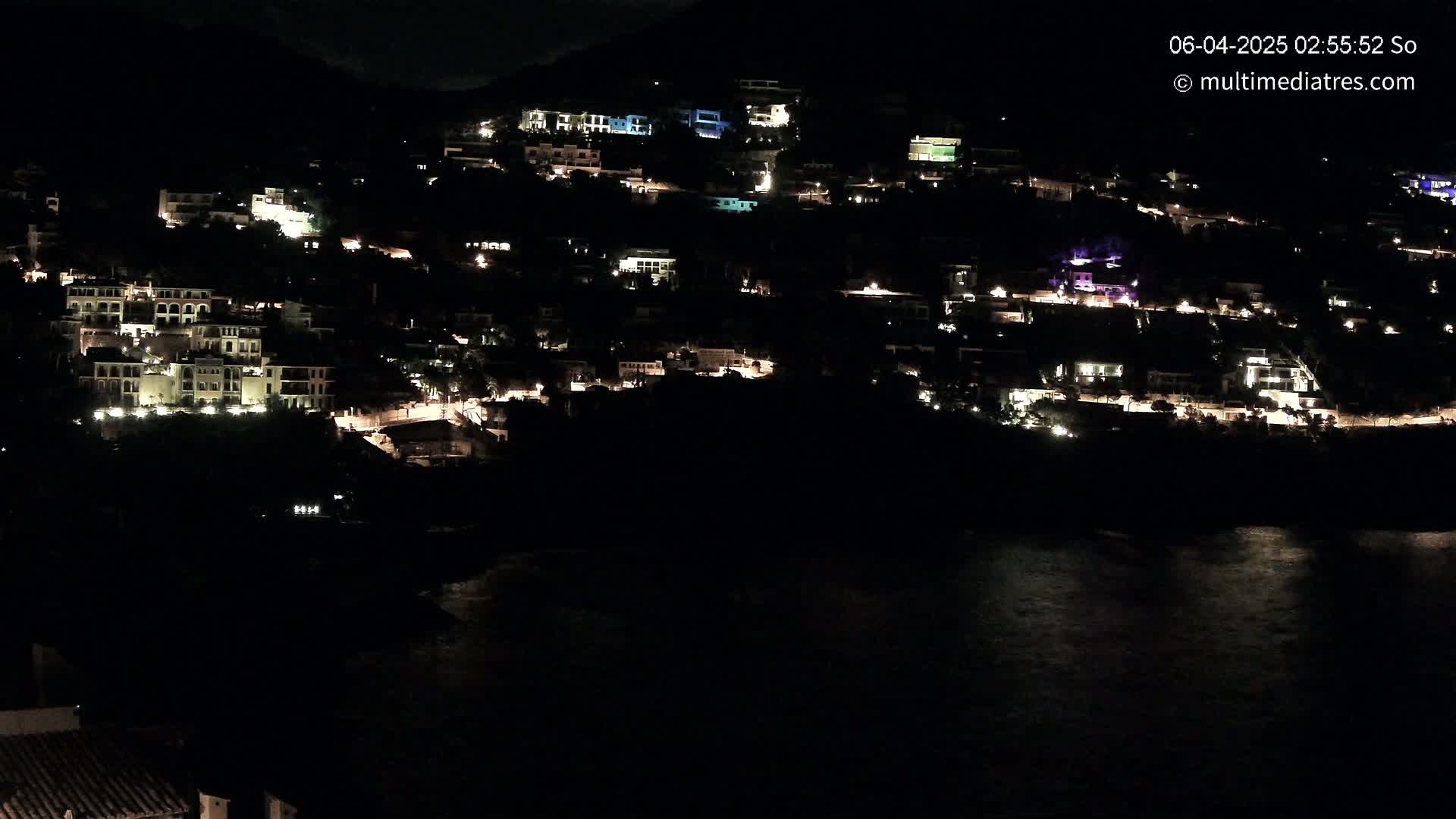 A hillside village's lights illuminate the nighttime scene overlooking dark water.