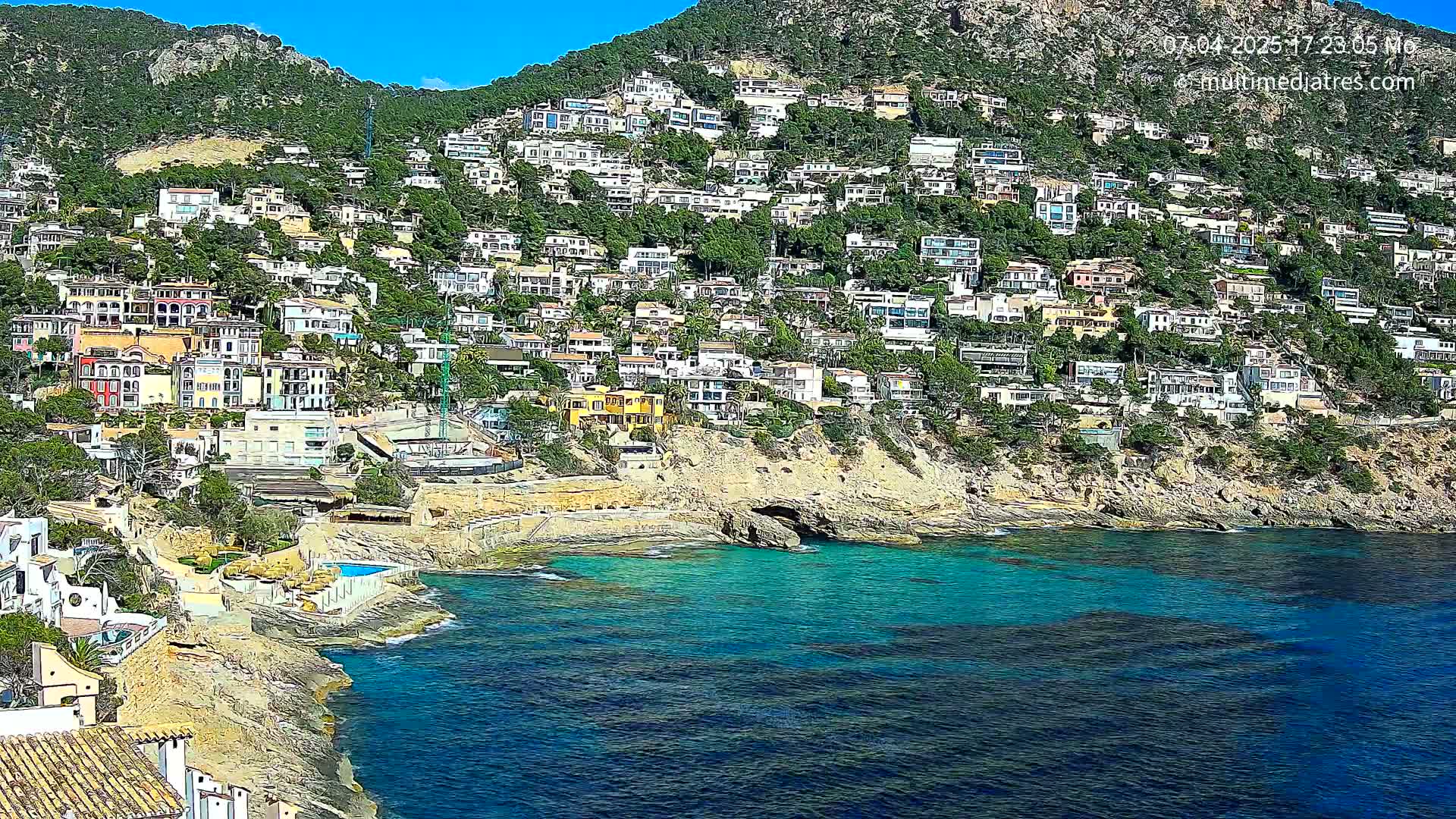 A hillside densely populated with houses slopes down to a clear, teal-colored ocean on a sunny day.