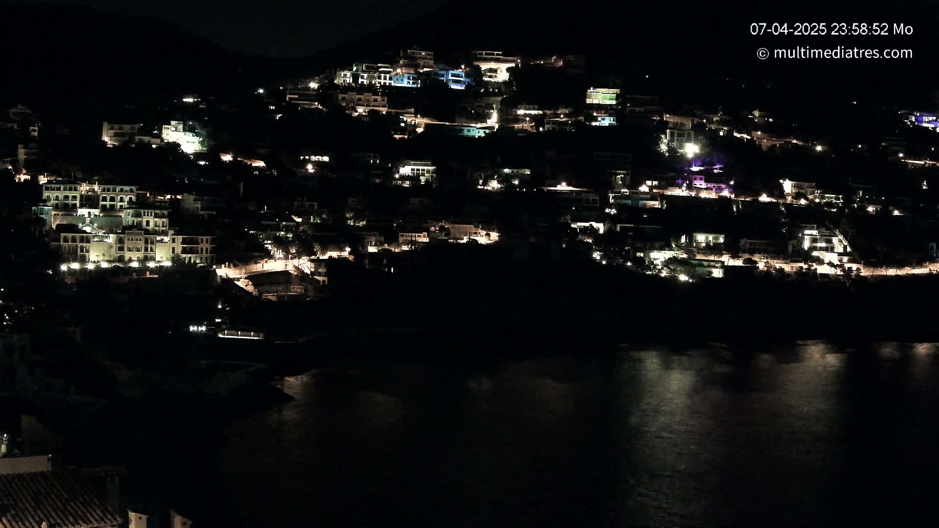 A hillside town is illuminated at night by the lights of numerous buildings, overlooking a dark body of water.