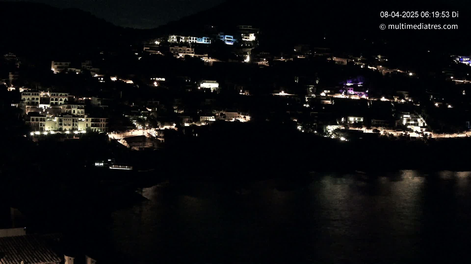 A hillside village is illuminated at night, with its lights reflected on the dark water below.