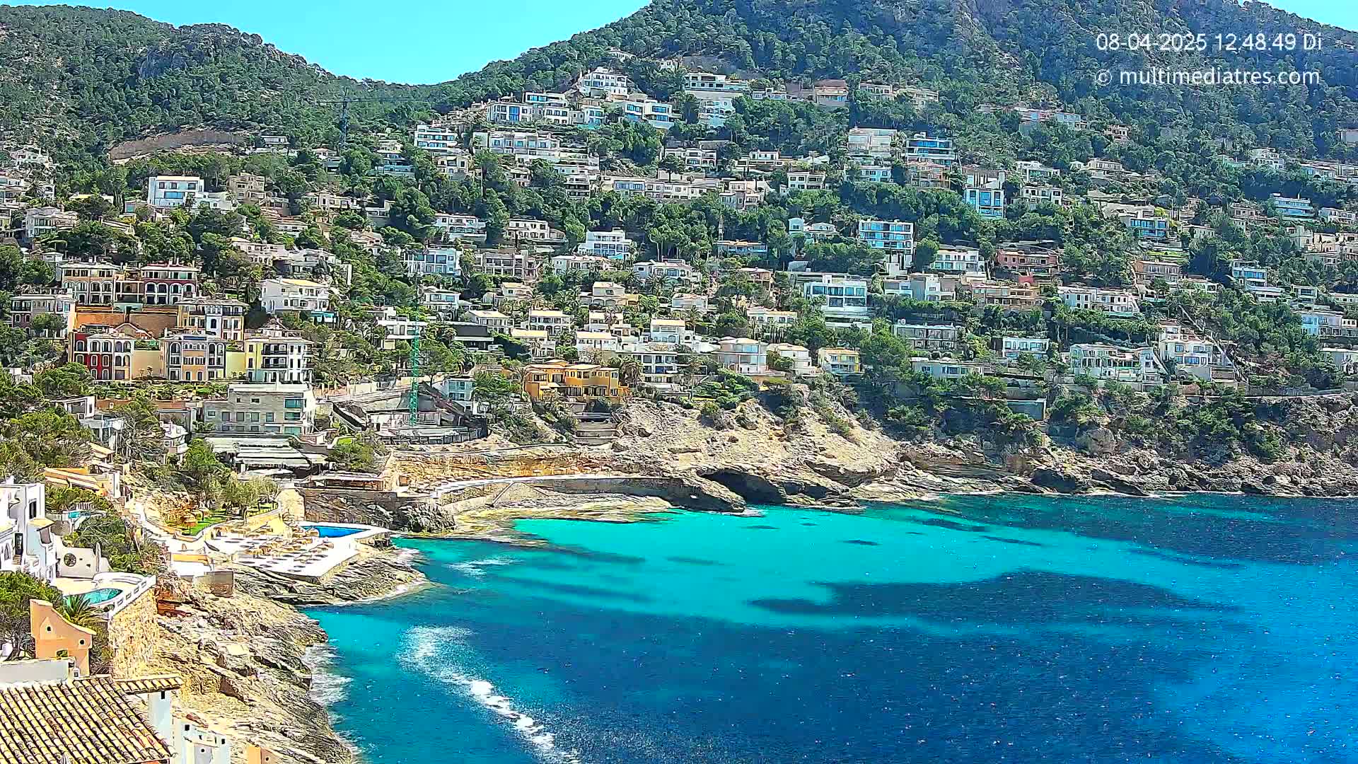 A hillside densely covered with houses overlooks a clear, turquoise bay on a sunny day.