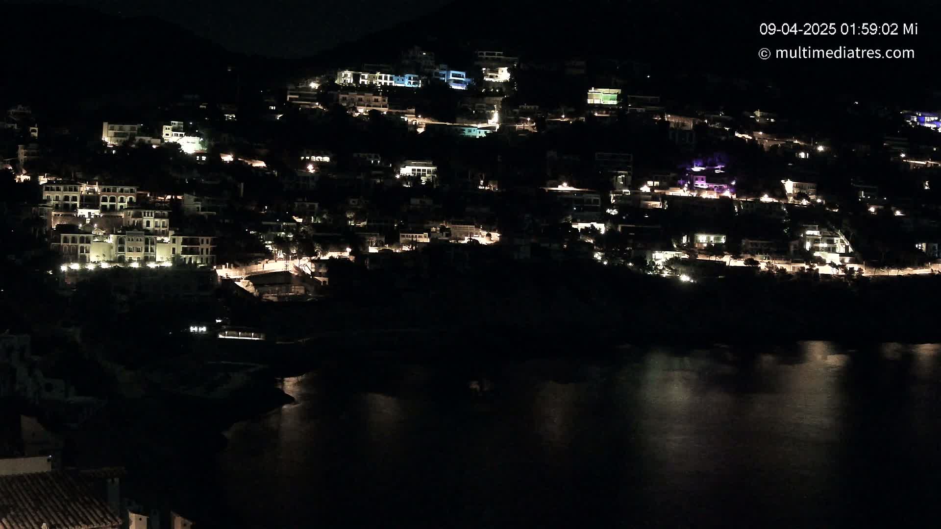 A hillside town at night, illuminated by numerous lights, overlooks a dark body of water under a clear, dark sky.