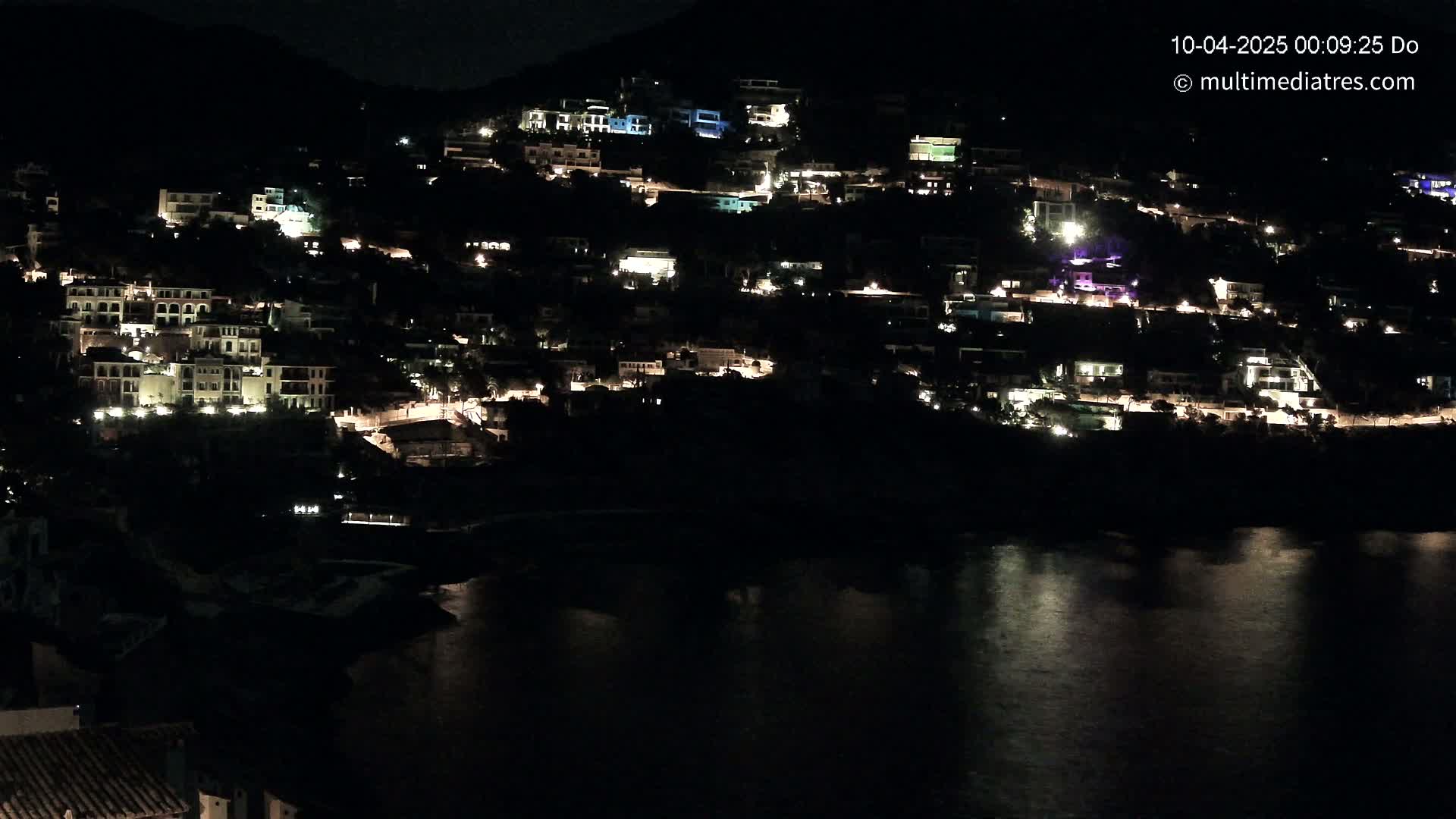 A hillside town's lights illuminate the night, overlooking a dark body of water reflecting the glow.