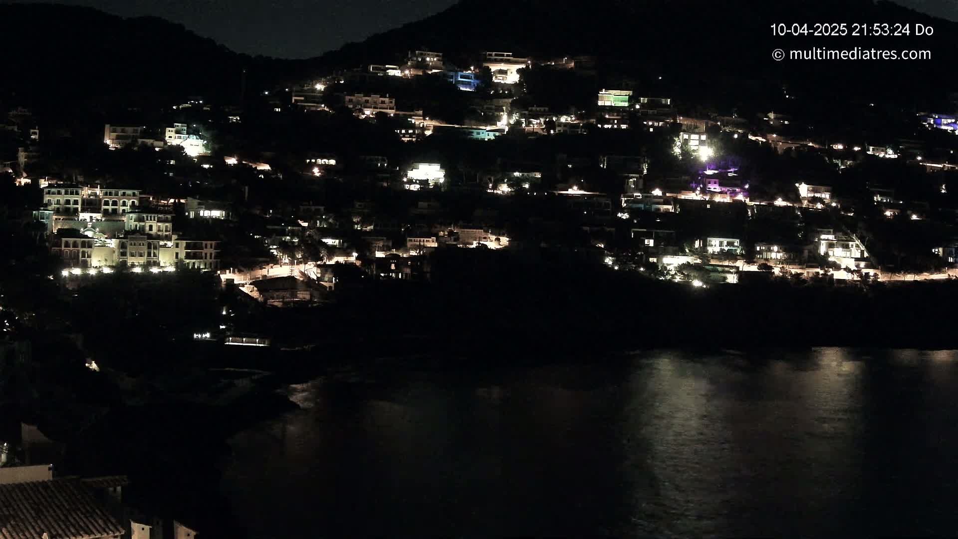 A hillside village is illuminated at night, its lights reflected in the calm water below.