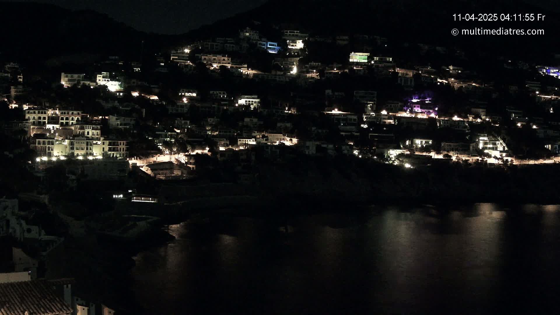 A hillside town with numerous illuminated buildings at night, overlooking a calm body of water.