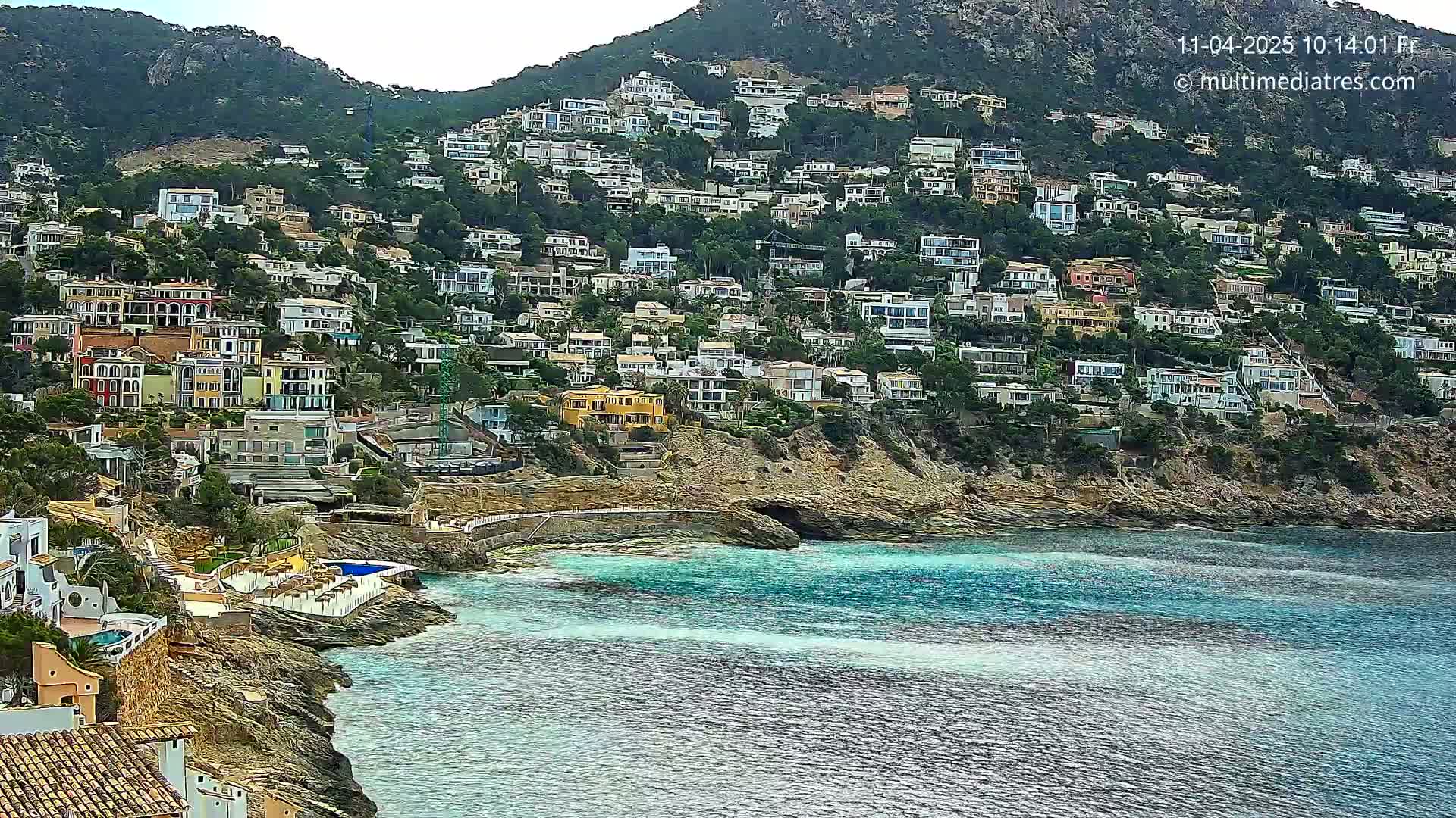 A hillside densely covered with houses slopes down to a calm, teal-colored sea.