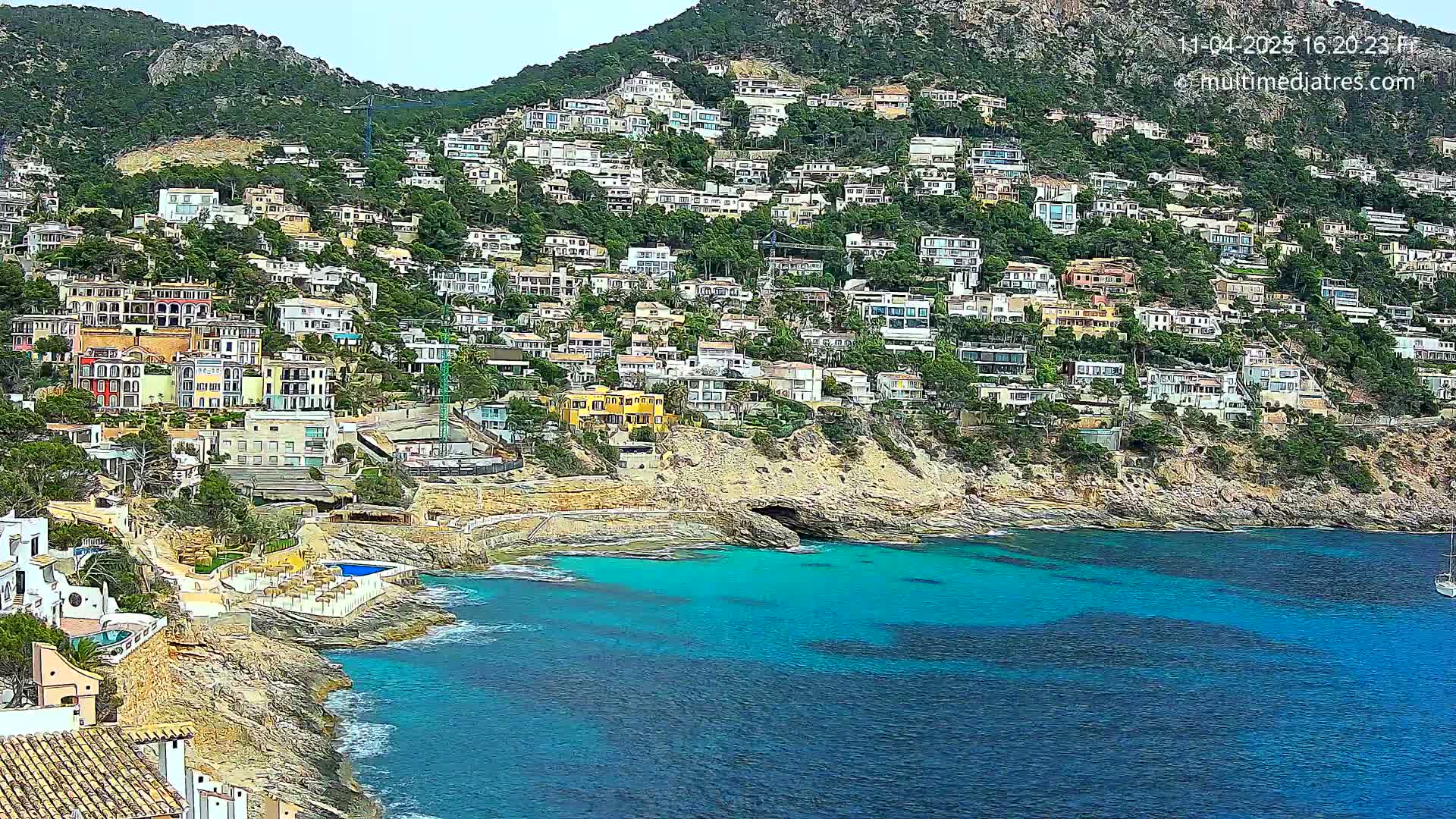 A hillside densely covered with houses overlooks a vibrant turquoise bay on a partly cloudy day.