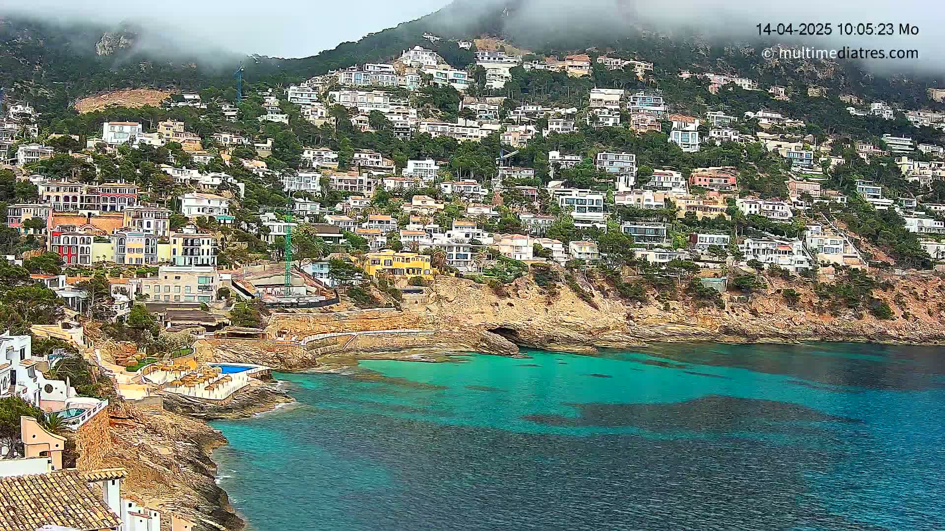 A hillside densely covered with houses overlooks a calm, teal-colored bay on a cloudy day.