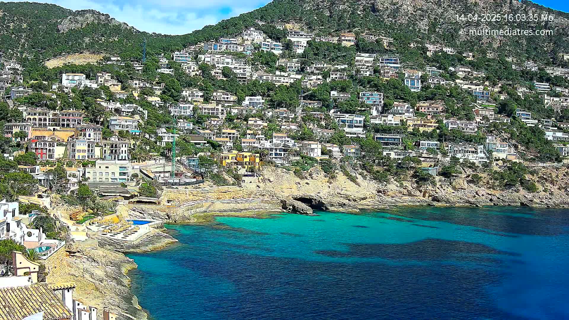 A hillside covered in houses slopes down to a rocky coast with clear, turquoise water under a partly sunny sky.