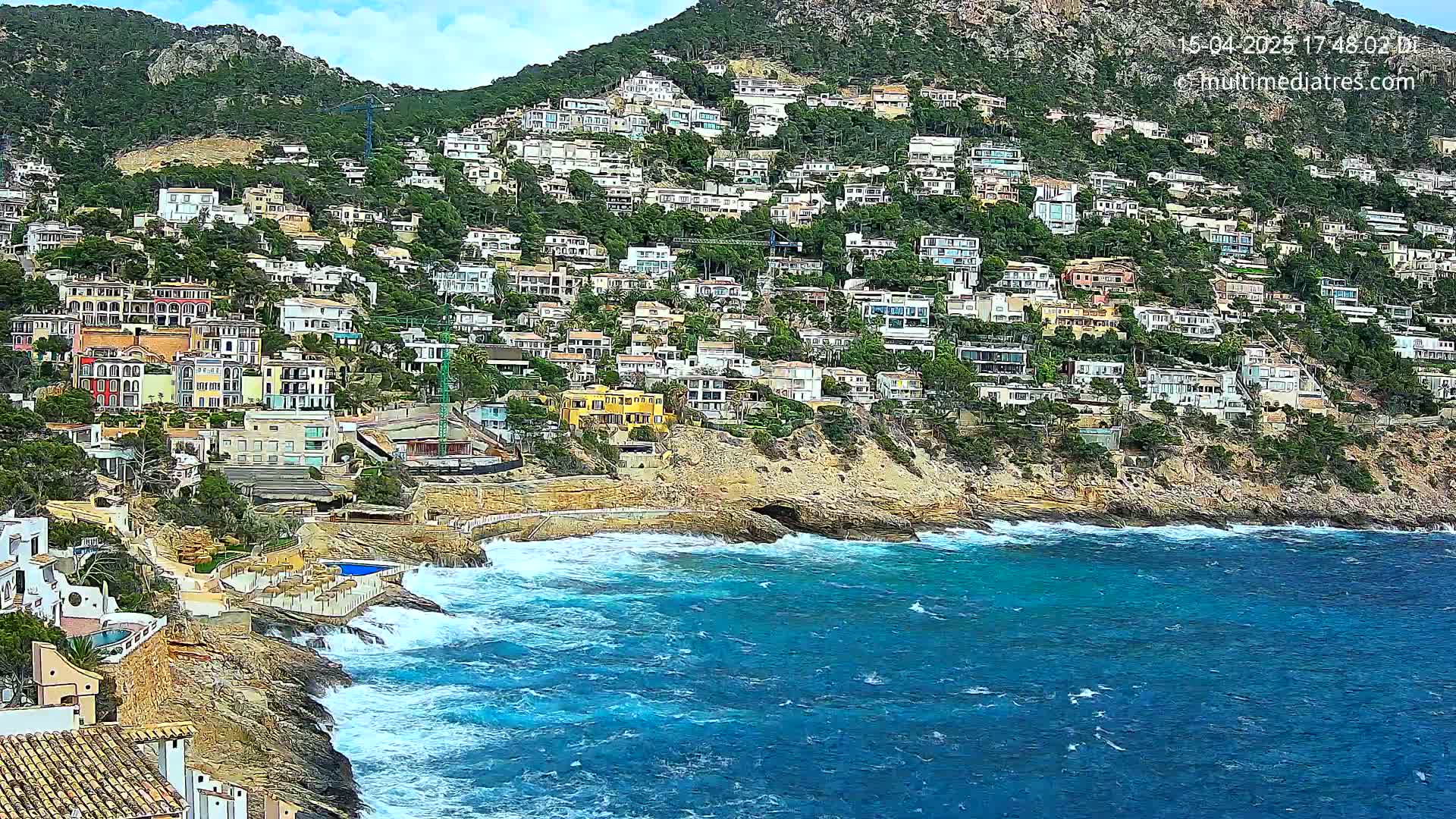 A hillside densely covered with houses overlooks a rocky coastline with a turbulent blue sea under a partly cloudy sky.
