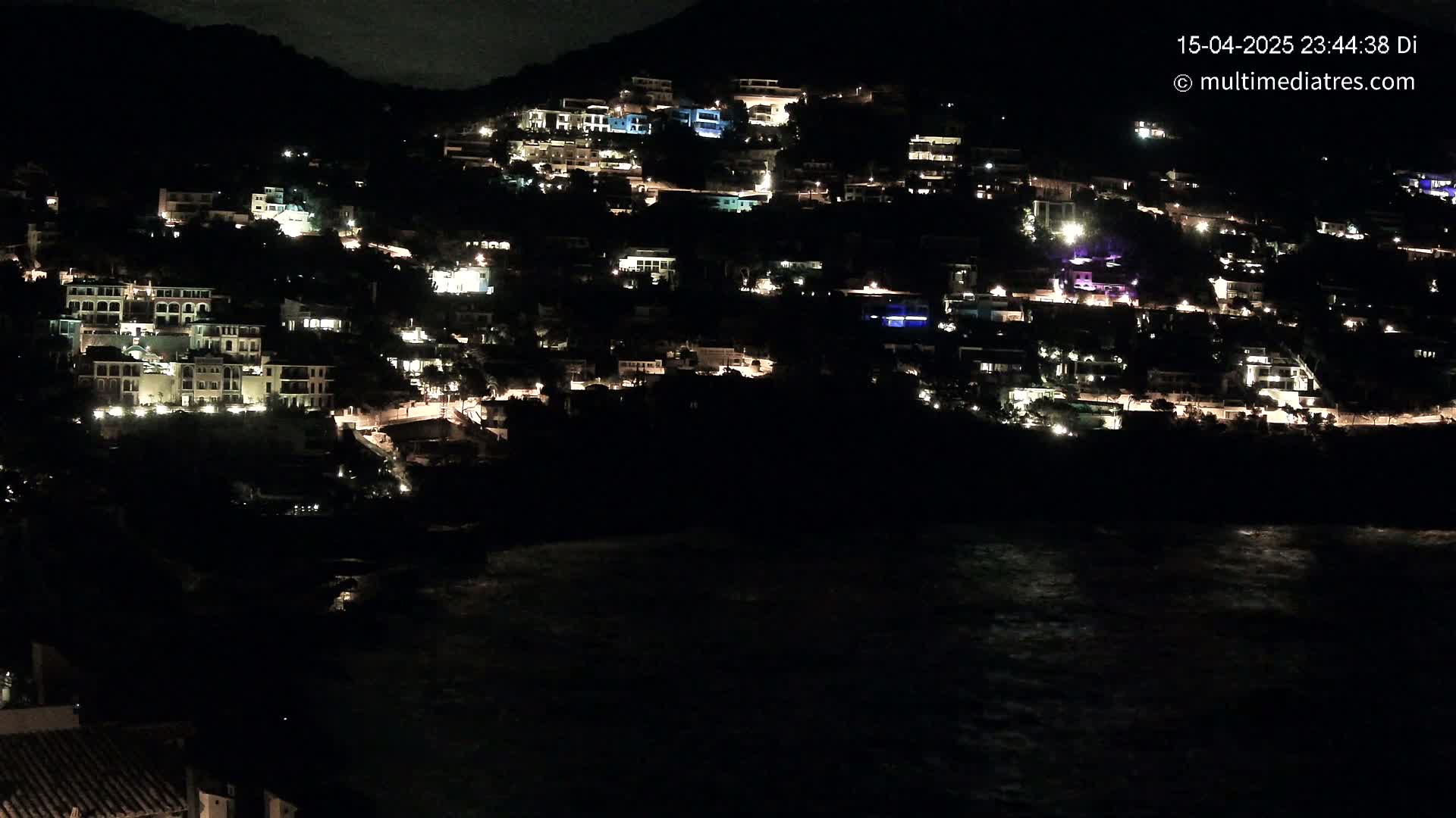 Many houses with illuminated windows are built on a dark hillside overlooking a dark, wavy body of water at night.