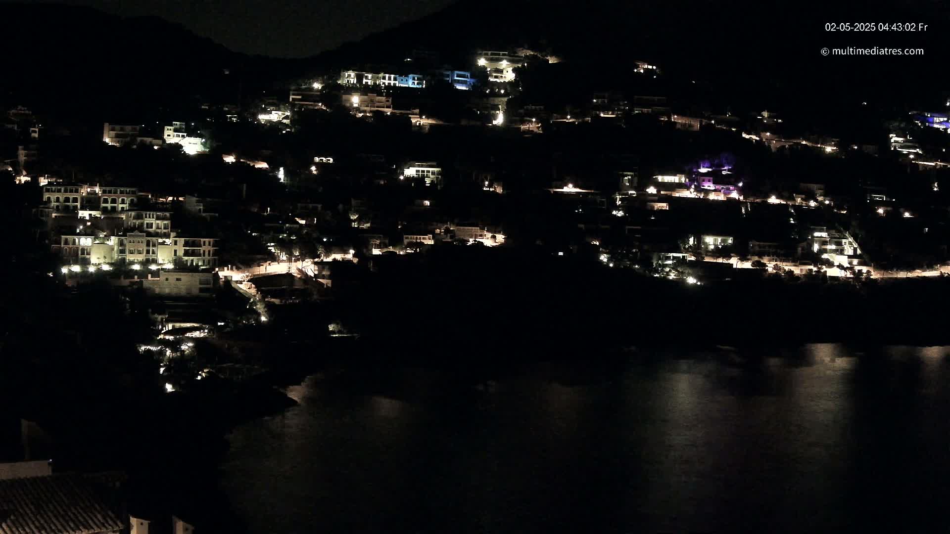 A hillside town is illuminated at night, its lights reflecting softly on a dark body of water below.