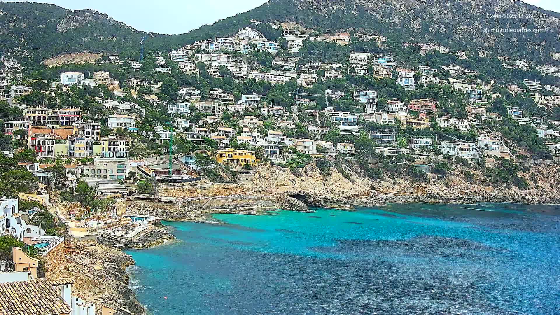 A hillside densely covered with houses overlooks a bright turquoise bay on a partly sunny day.