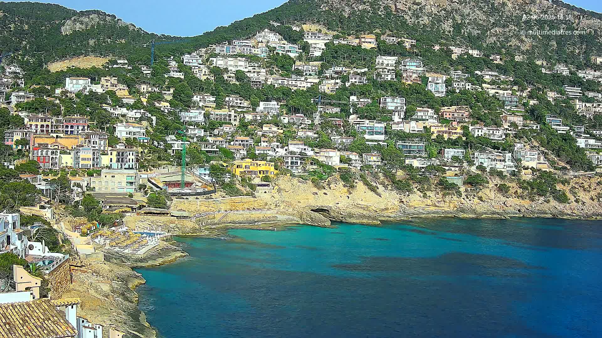 A hillside covered in houses slopes down to a clear, turquoise cove on a sunny day.