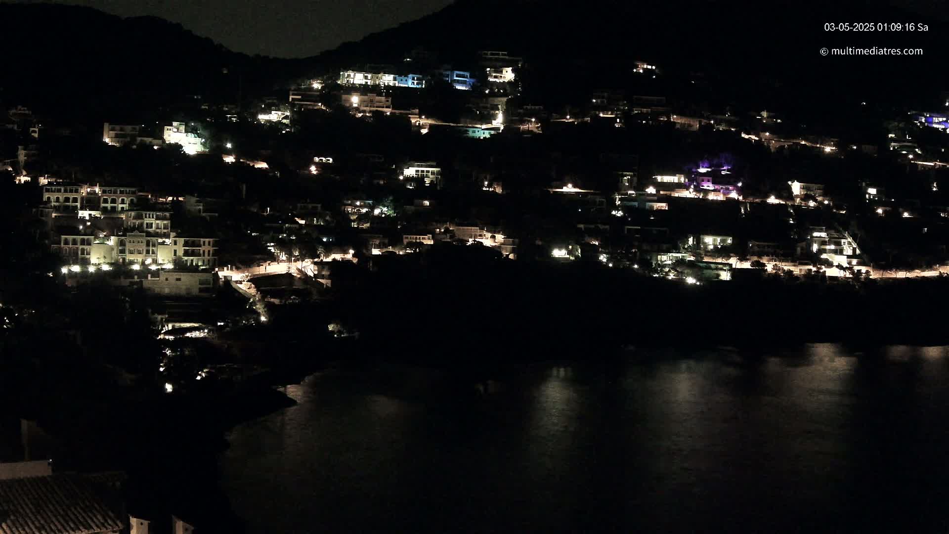 A hillside village is illuminated at night by the lights of many houses, overlooking a dark body of water.