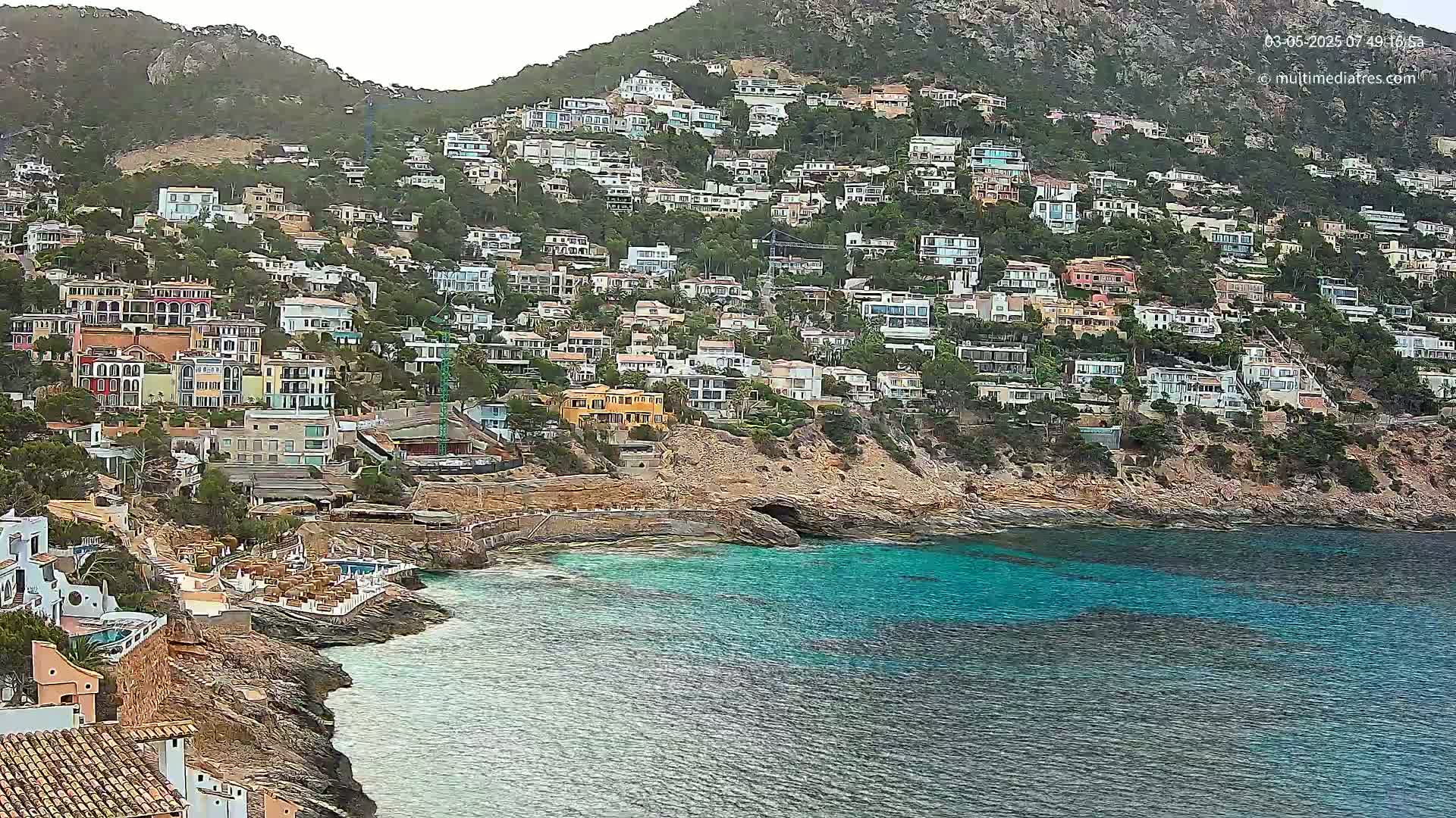 A hillside covered in houses slopes down to a calm, teal-colored ocean.