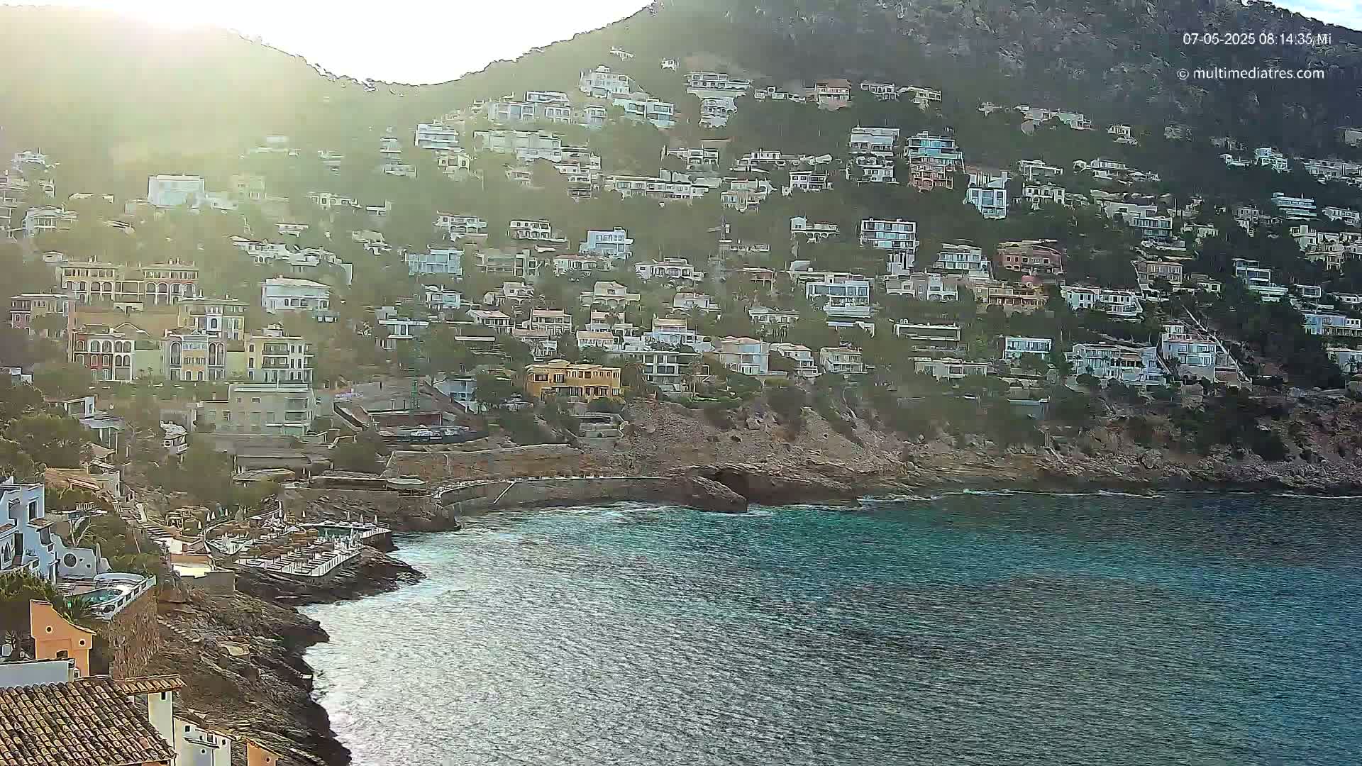 A hillside densely covered with houses slopes down to a rocky coastline and calm, blue-green water under a hazy, sunny sky.
