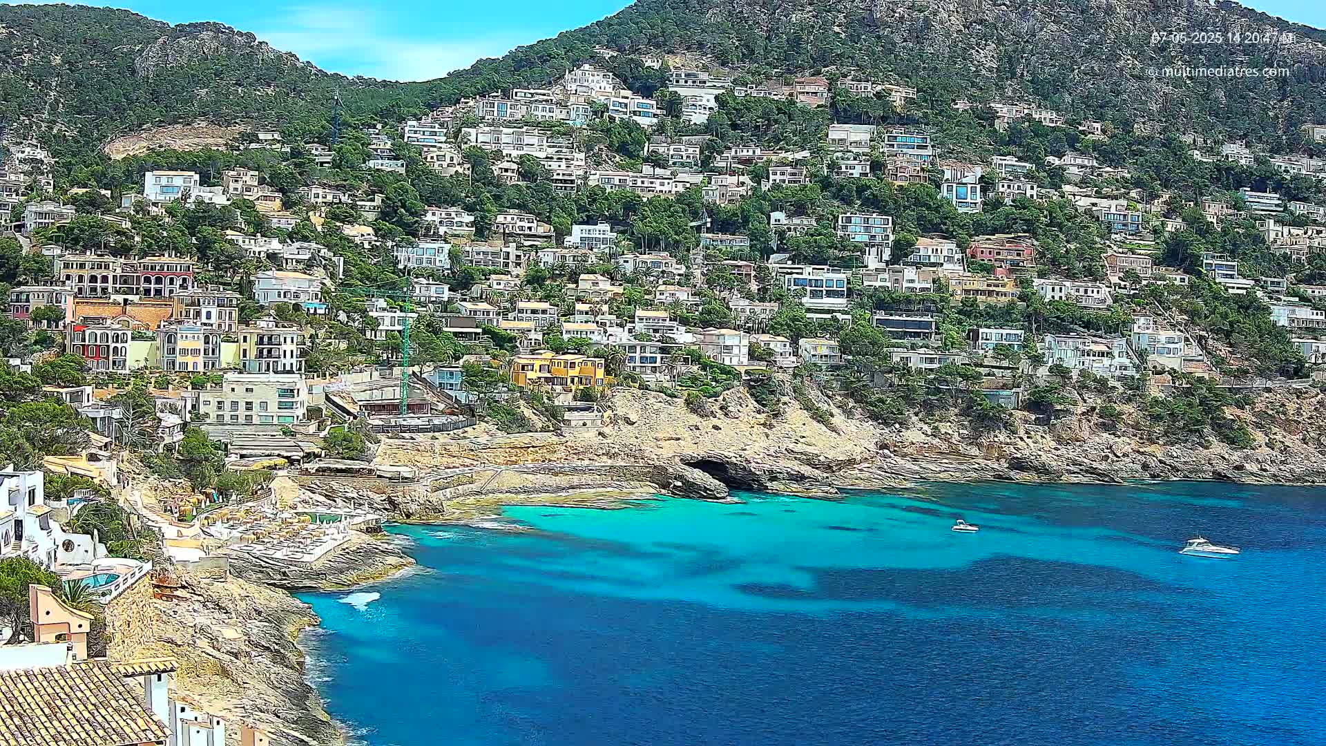 A hillside densely covered with houses overlooks a clear, turquoise bay where two boats are visible on a sunny day.