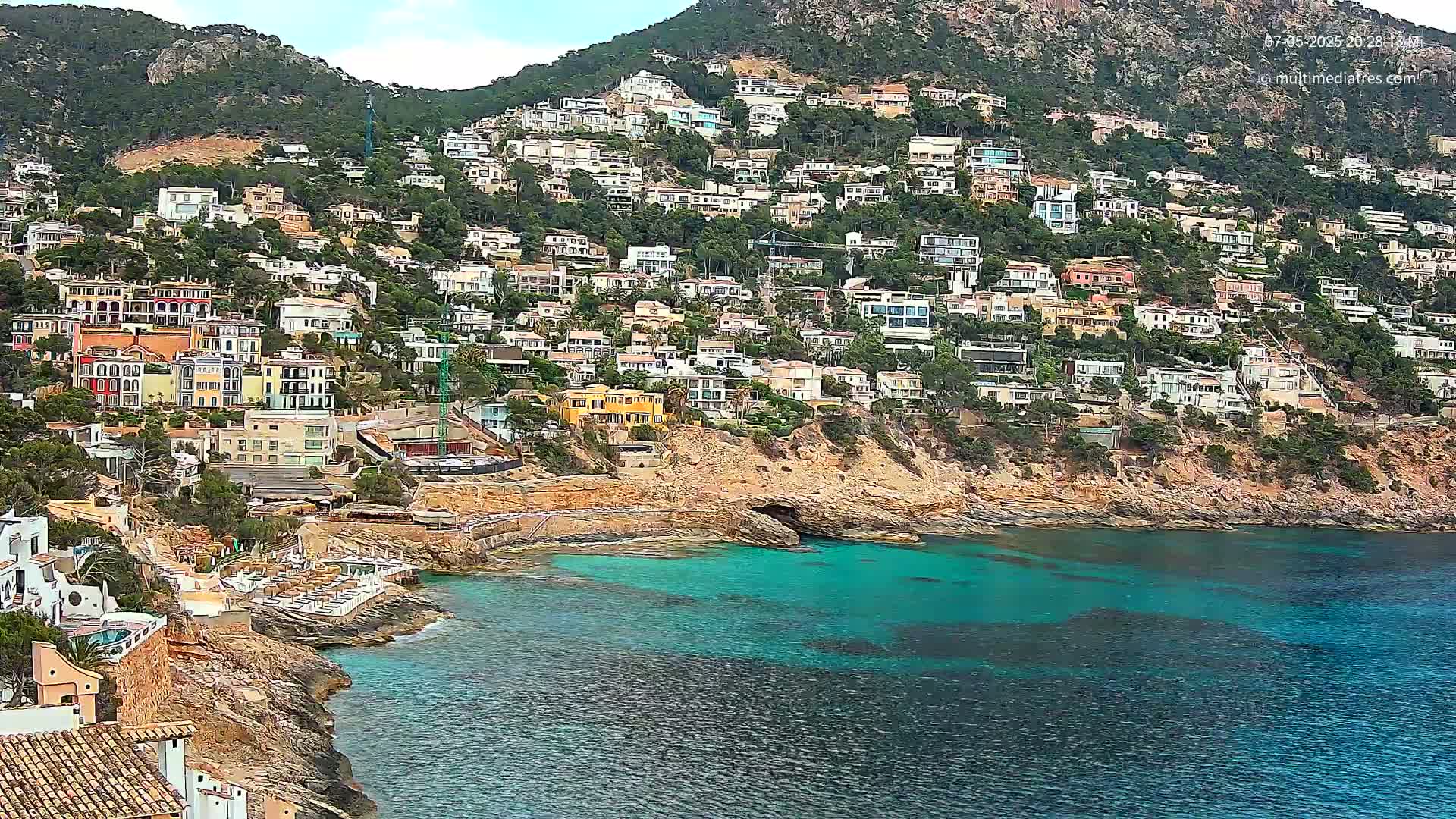 A hillside town of colorful houses clings to a rocky coast, overlooking a calm, turquoise bay under a partly cloudy sky.