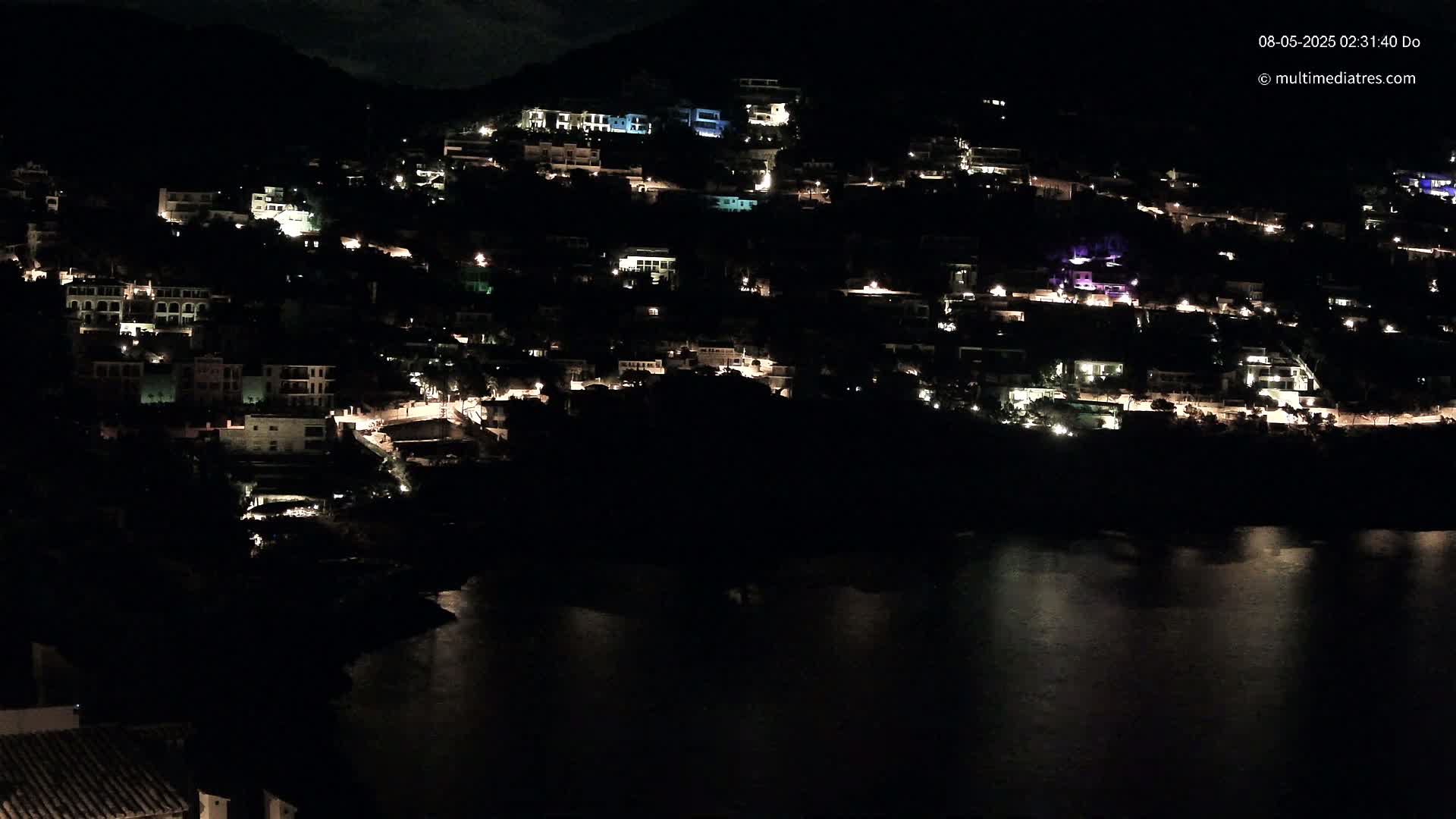 A hillside town at night, with many houses lit up, overlooks a dark body of water under a mostly clear sky.
