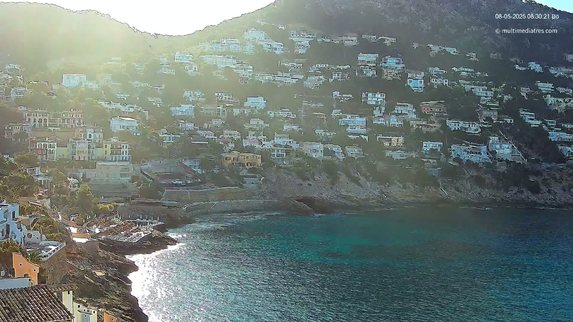 A hillside covered in houses slopes down to a rocky cove with clear turquoise water and gentle waves under a hazy, sunny sky.