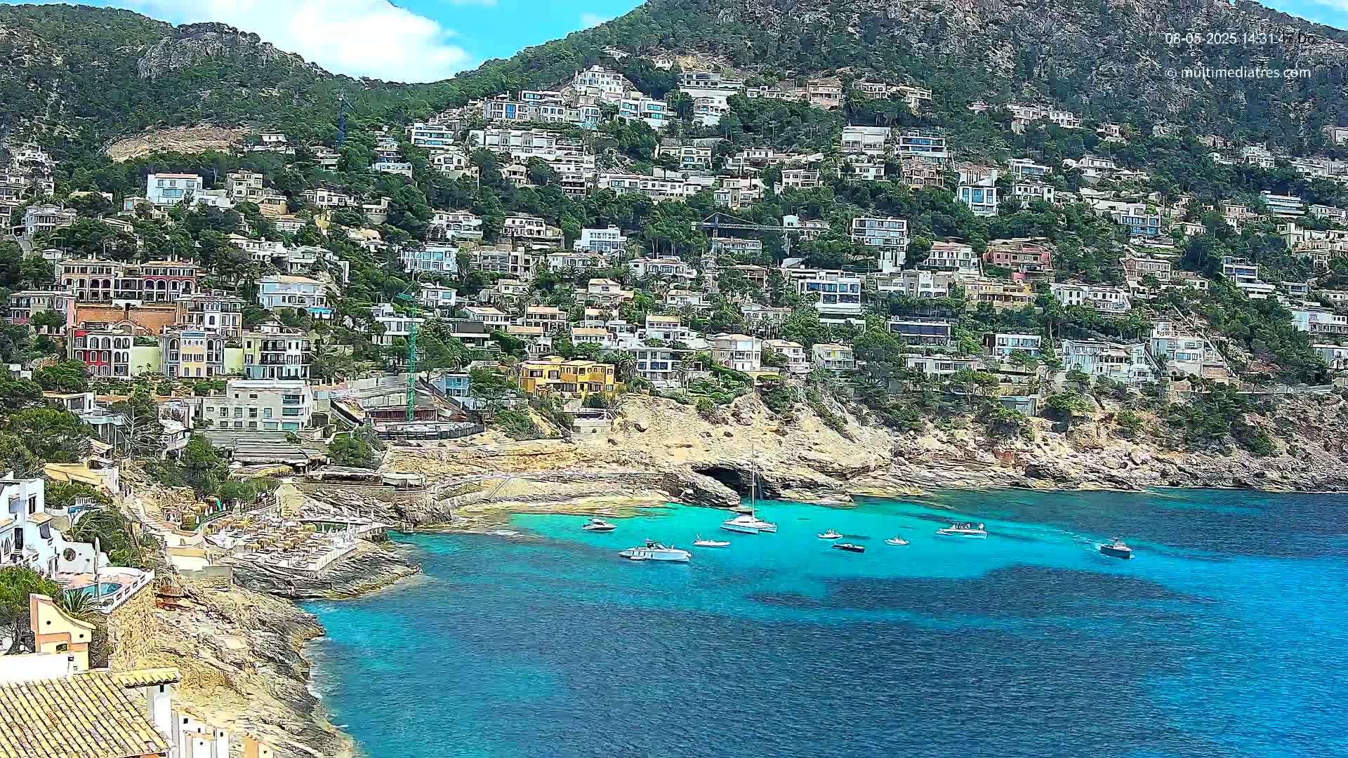 A hillside covered in houses slopes down to a calm, turquoise cove with several boats anchored in the clear water under a partly sunny sky.