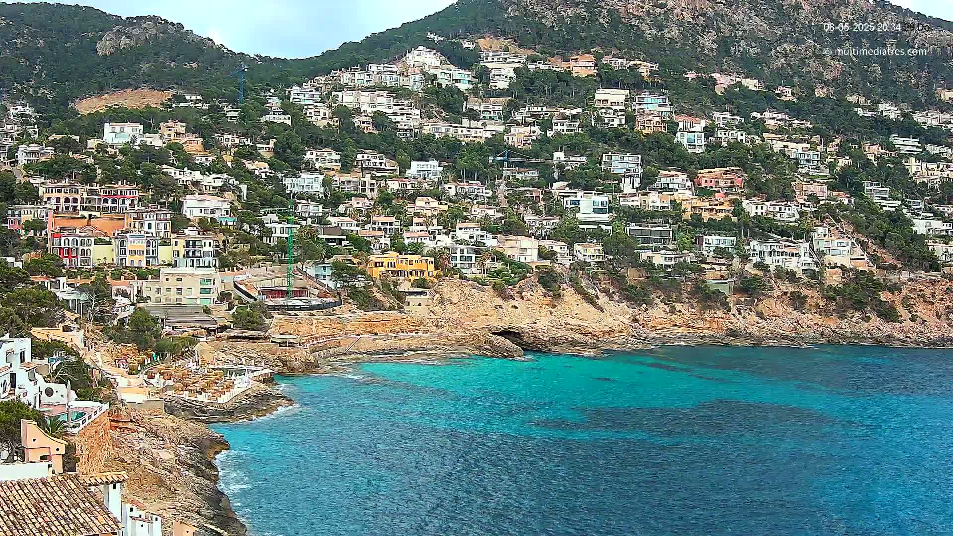 A hillside covered in houses slopes down to a turquoise bay on an overcast day.