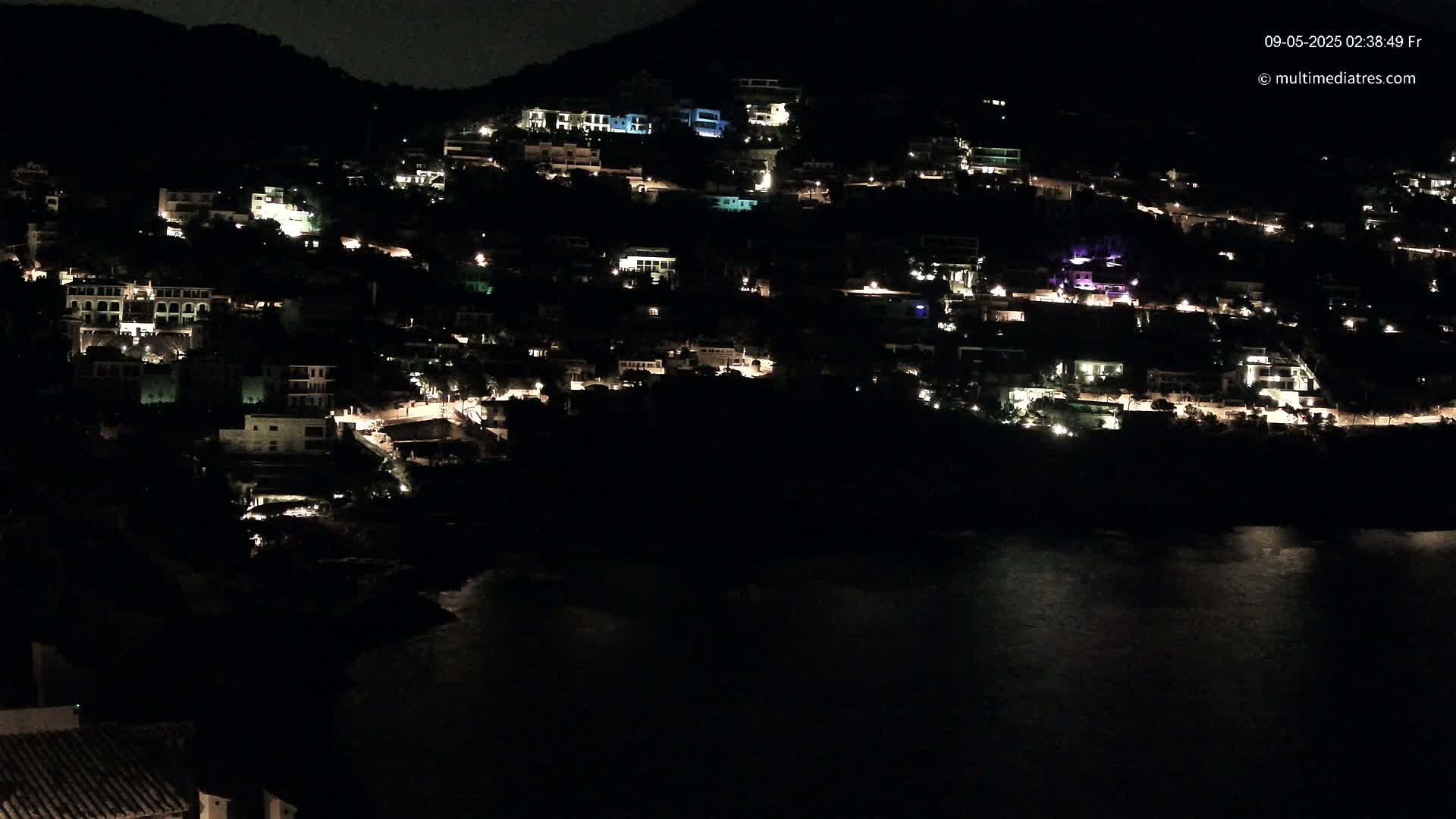 A hillside town is illuminated at night, its lights scattered across the dark landscape next to a calm body of water.