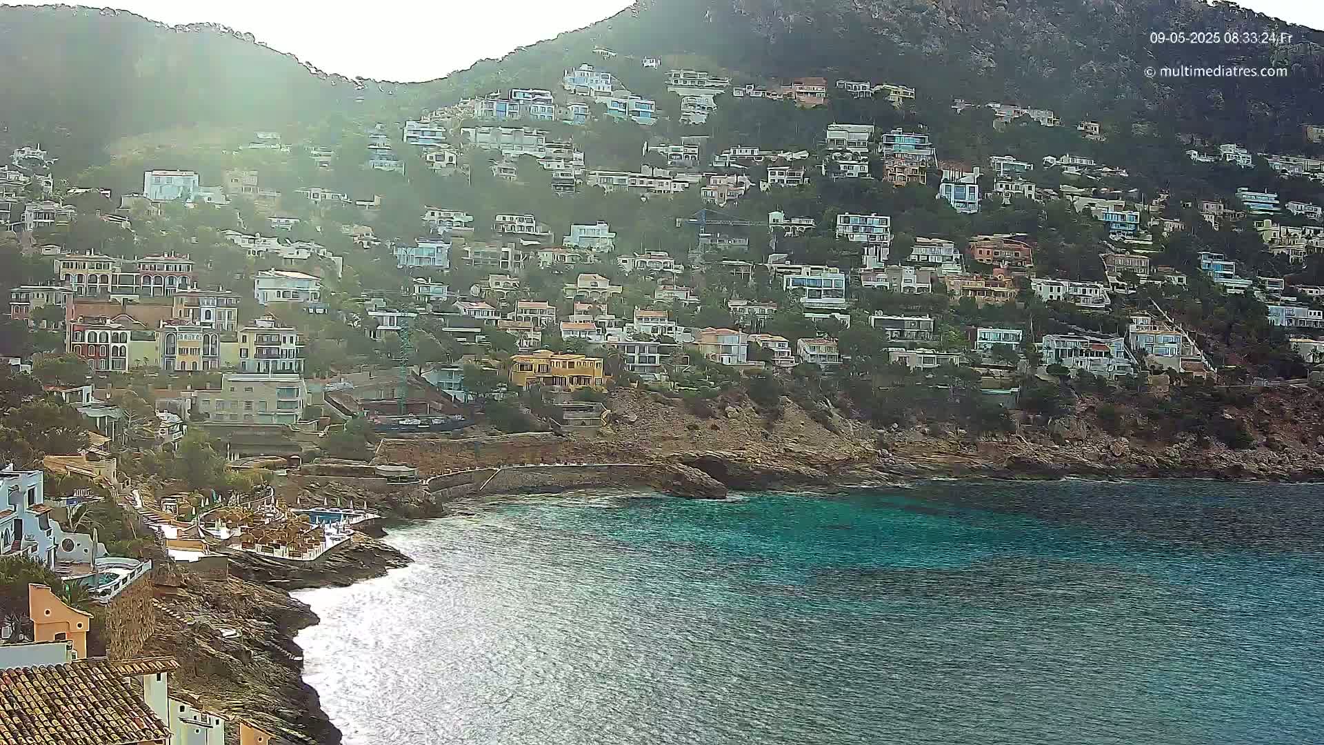 A hillside covered in houses overlooks a calm, teal ocean on a hazy day.