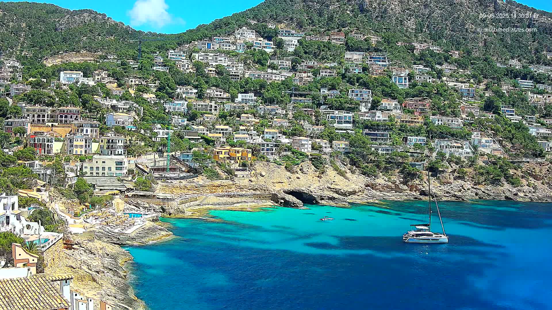 A hillside densely covered with houses overlooks a clear, turquoise bay where a sailboat is anchored under a bright, sunny sky.