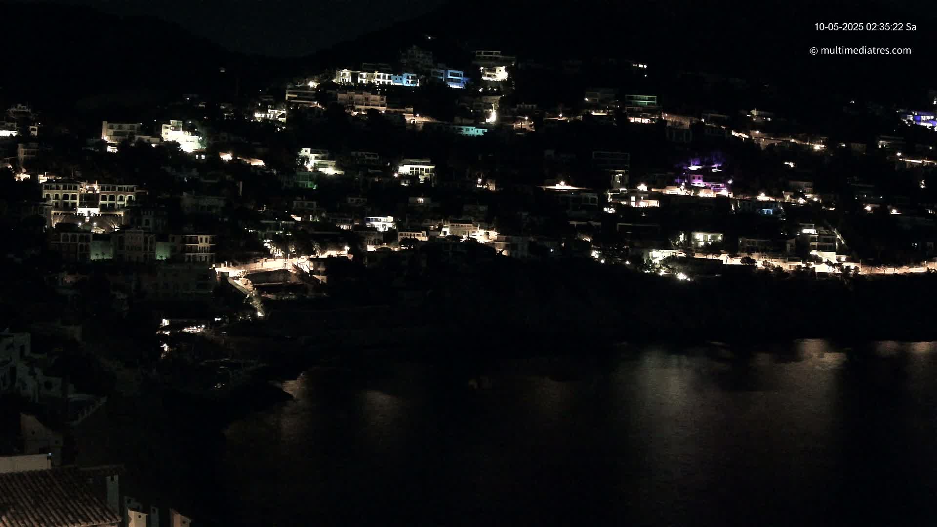 A hillside town at night, with many houses lit up, overlooks a dark body of water under a clear, dark sky.