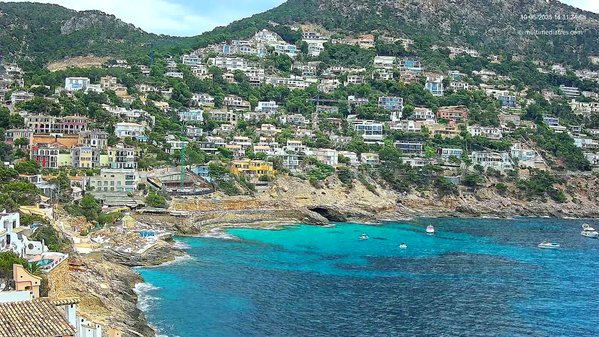A hillside densely covered with houses overlooks a calm, turquoise bay with a few small boats.