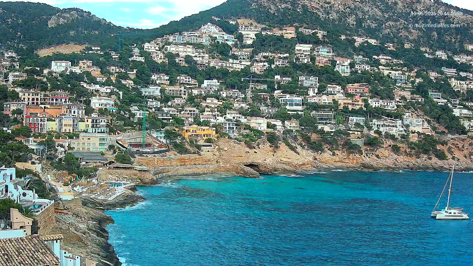 A hillside covered in houses overlooks a calm, turquoise bay with a sailboat anchored in the distance under a partly cloudy sky.