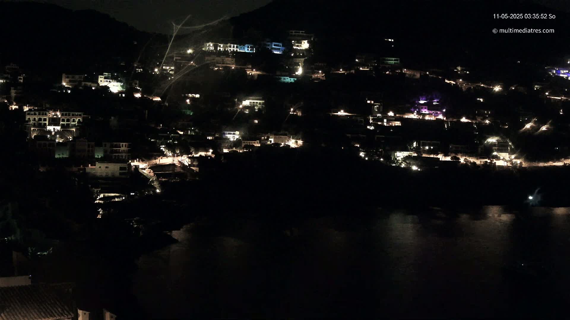 A hillside town's lights illuminate the night, overlooking a dark body of water under a clear, starless sky.