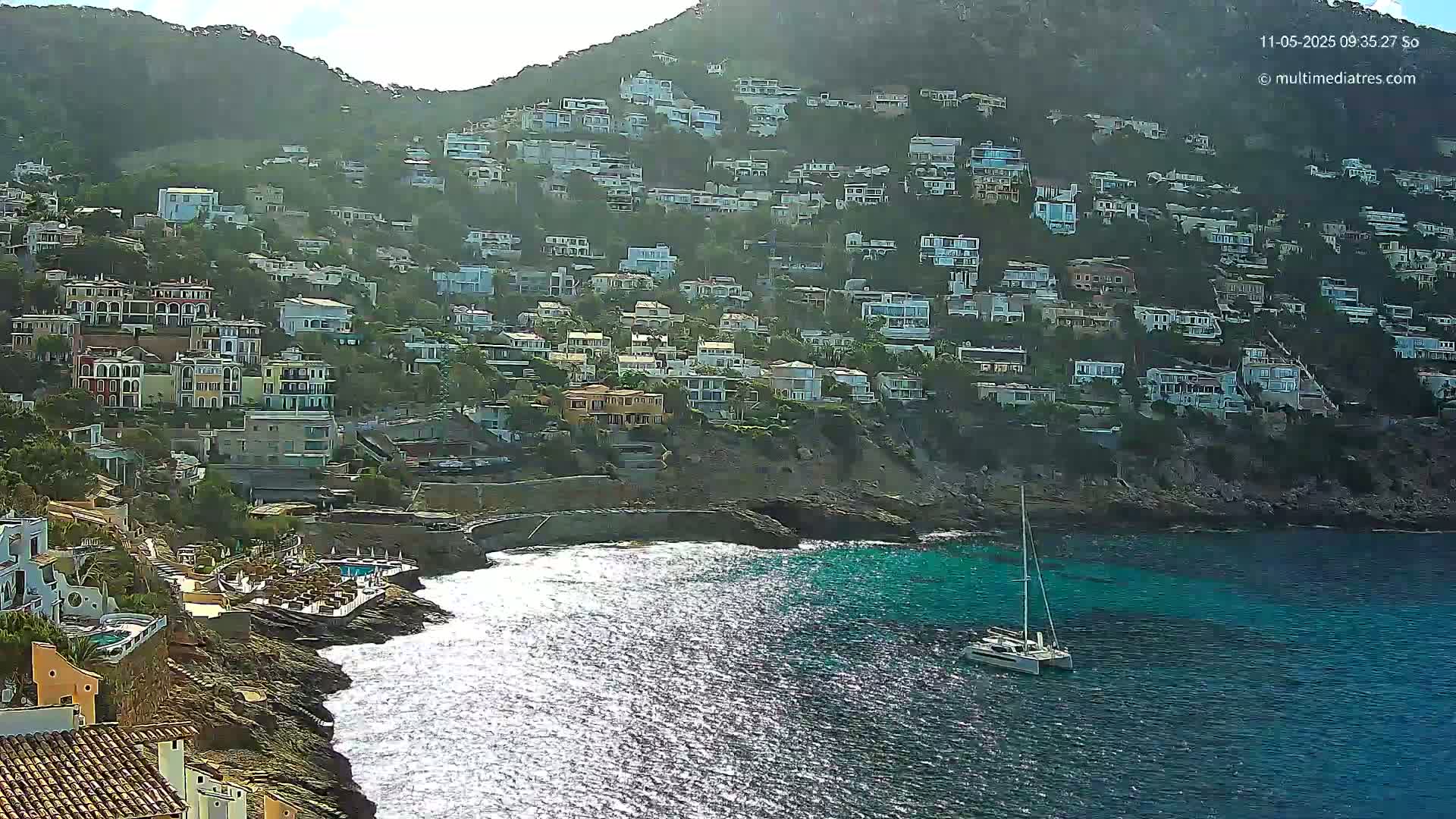 A hillside covered in houses slopes down to a rocky coast where a sailboat is anchored in bright, sunny conditions with somewhat choppy water.